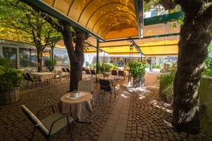 Un área de patio al aire libre con mesas y sillas, cubierta por un toldo amarillo, rodeada de árboles y plantas en macetas.