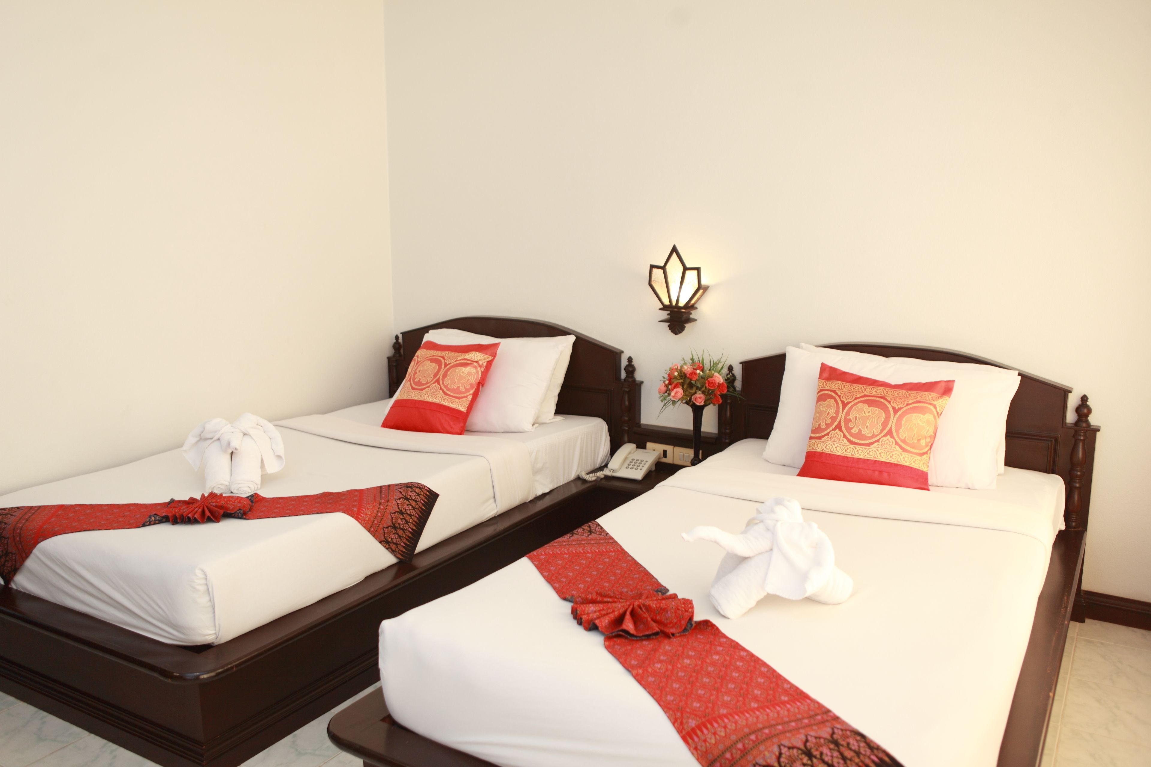 Habitación de hotel con dos camas, cada una con ropa de cama blanca y almohadas decorativas rojas. Hay un aplique sobre las camas y una mesita de noche con un jarrón de flores entre ellas.