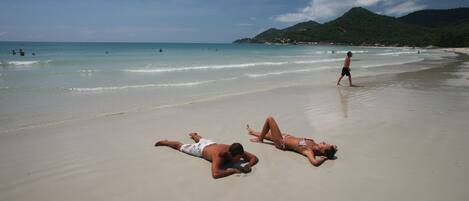 A beach with two people sunbathing and a person walking in the background.