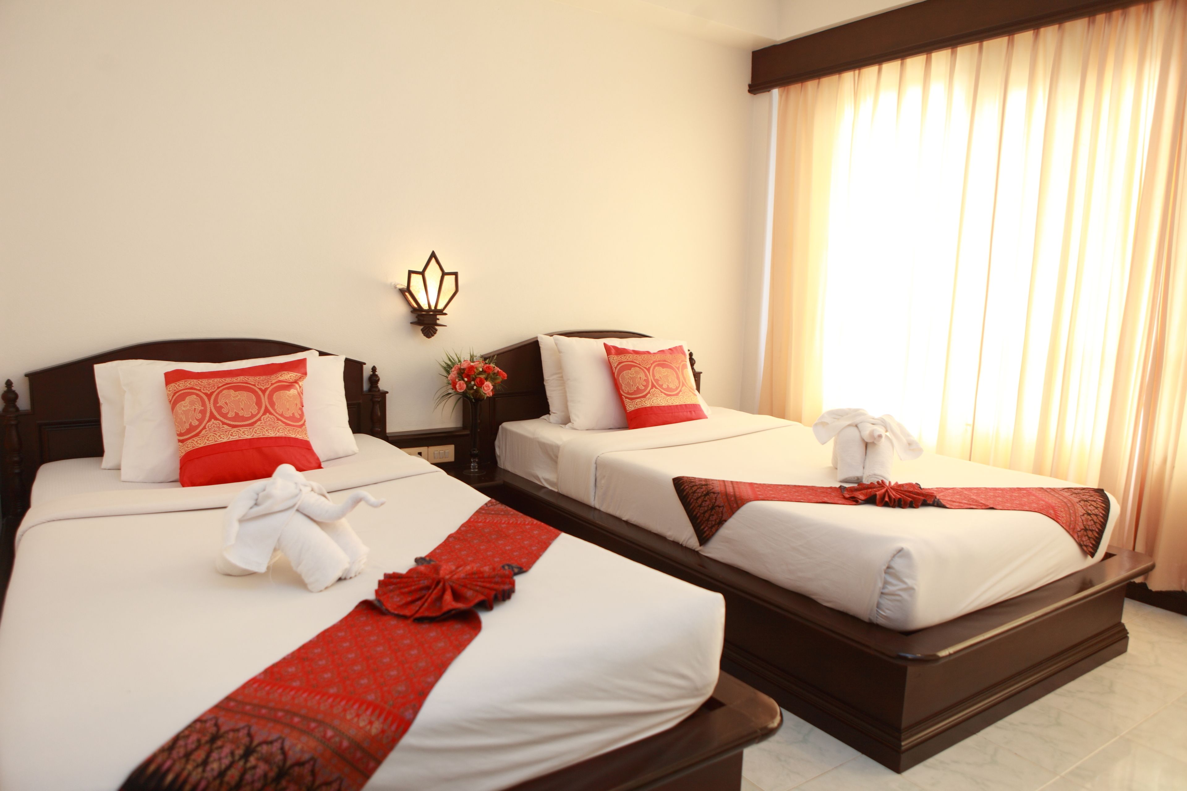 Dos camas contiguas con ropa de cama blanca y detalles rojos, un aplique en la pared y una ventana con cortinas.