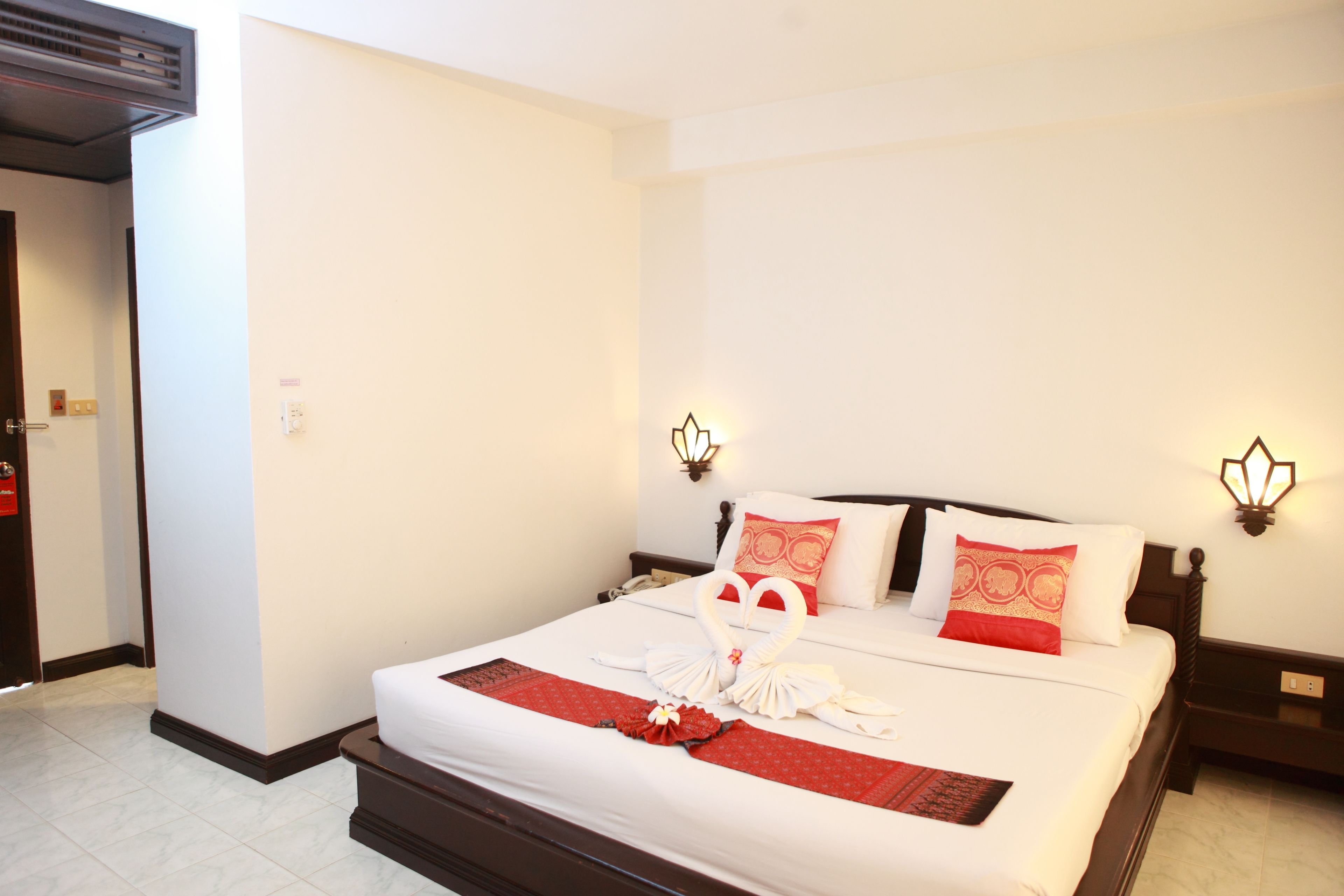 A hotel room with a neatly made bed, a red and white pillow, and a wall-mounted light fixture.