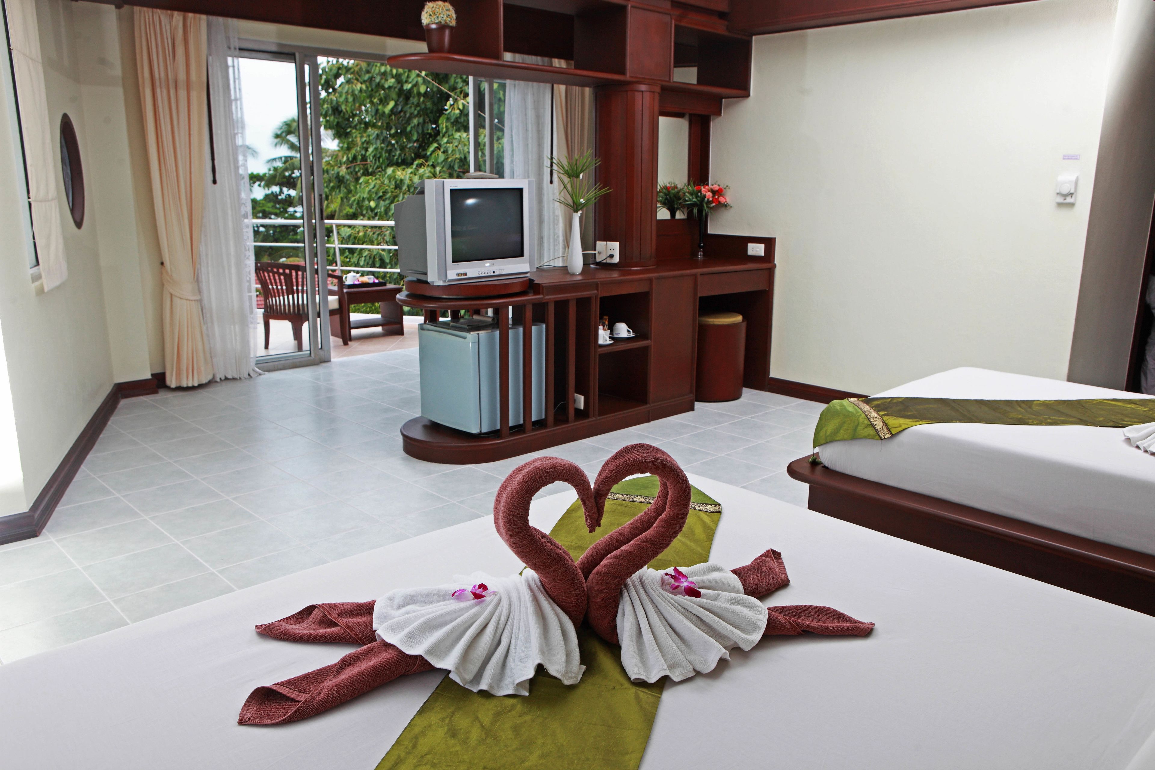 A hotel room with two beds, a TV, a desk, and a balcony with a view of trees.