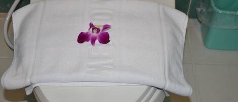 A white toilet with a white towel covering the seat and a purple flower embroidered on it.