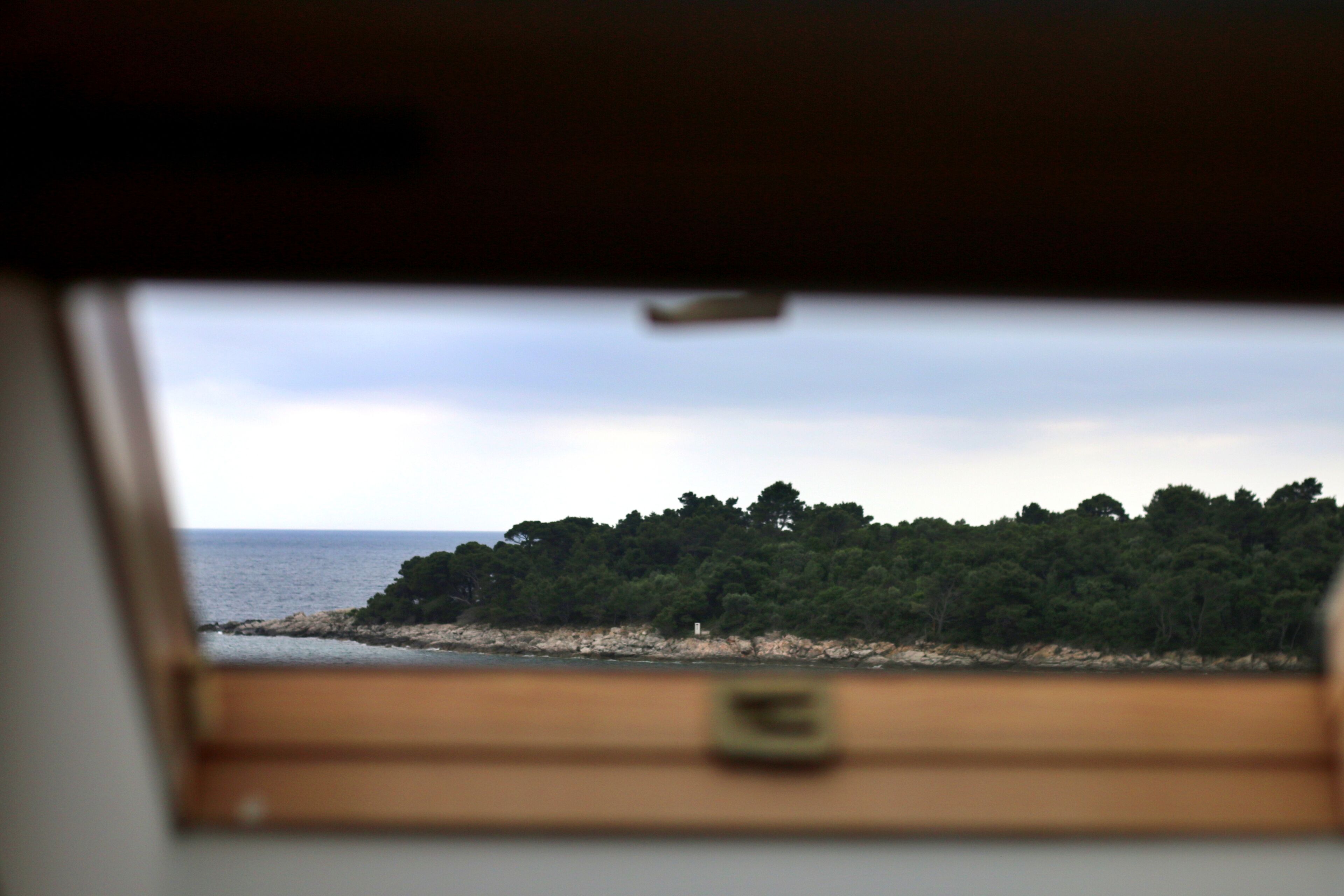 Blick aus einem Fahrzeug durch eine Fensterscheibe auf einen Strand, ein bewaldetes Ufer und das Meer in der Ferne.