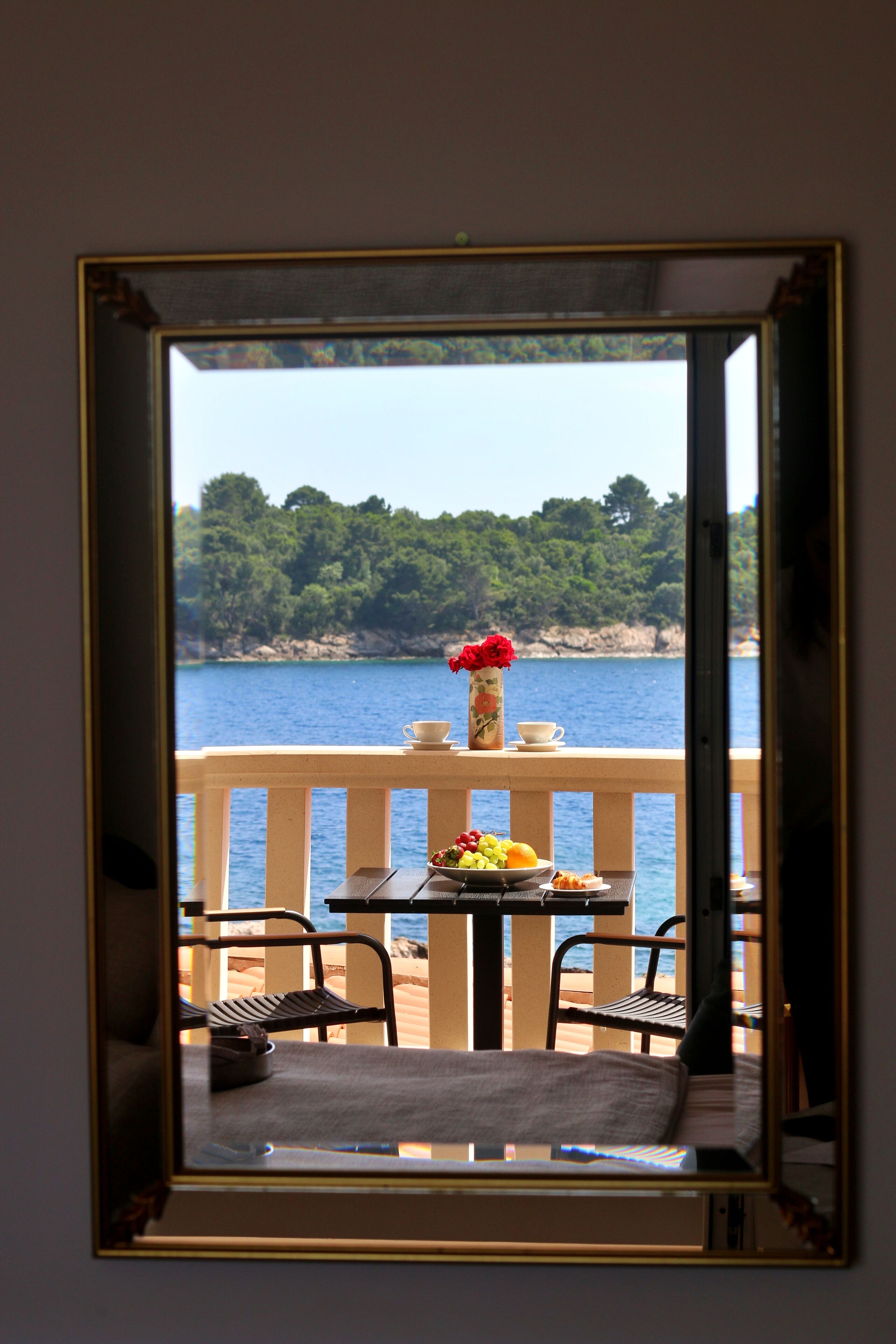 A balcony with a table set for two, a vase with flowers, and a view of a body of water and greenery.