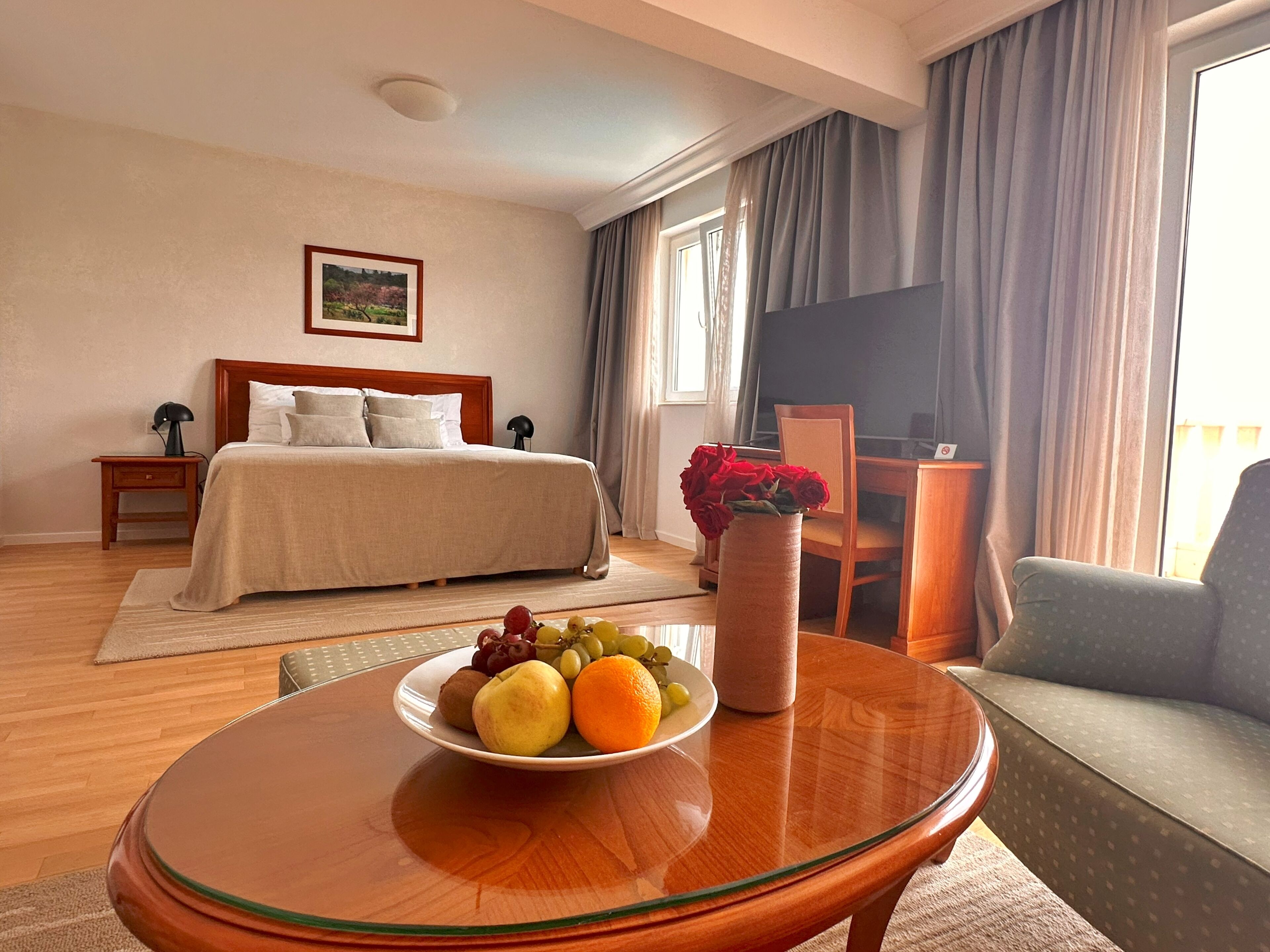 Quarto de hotel com uma cama, uma escrivaninha com cadeira, uma mesa redonda de vidro com uma fruteira e uma televisão na parede.