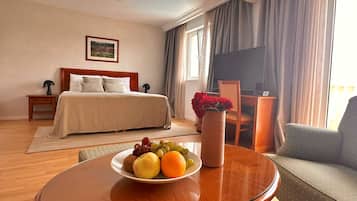 Quarto de hotel com uma cama, uma escrivaninha com cadeira, uma mesa redonda de vidro com uma fruteira e uma televisão na parede.