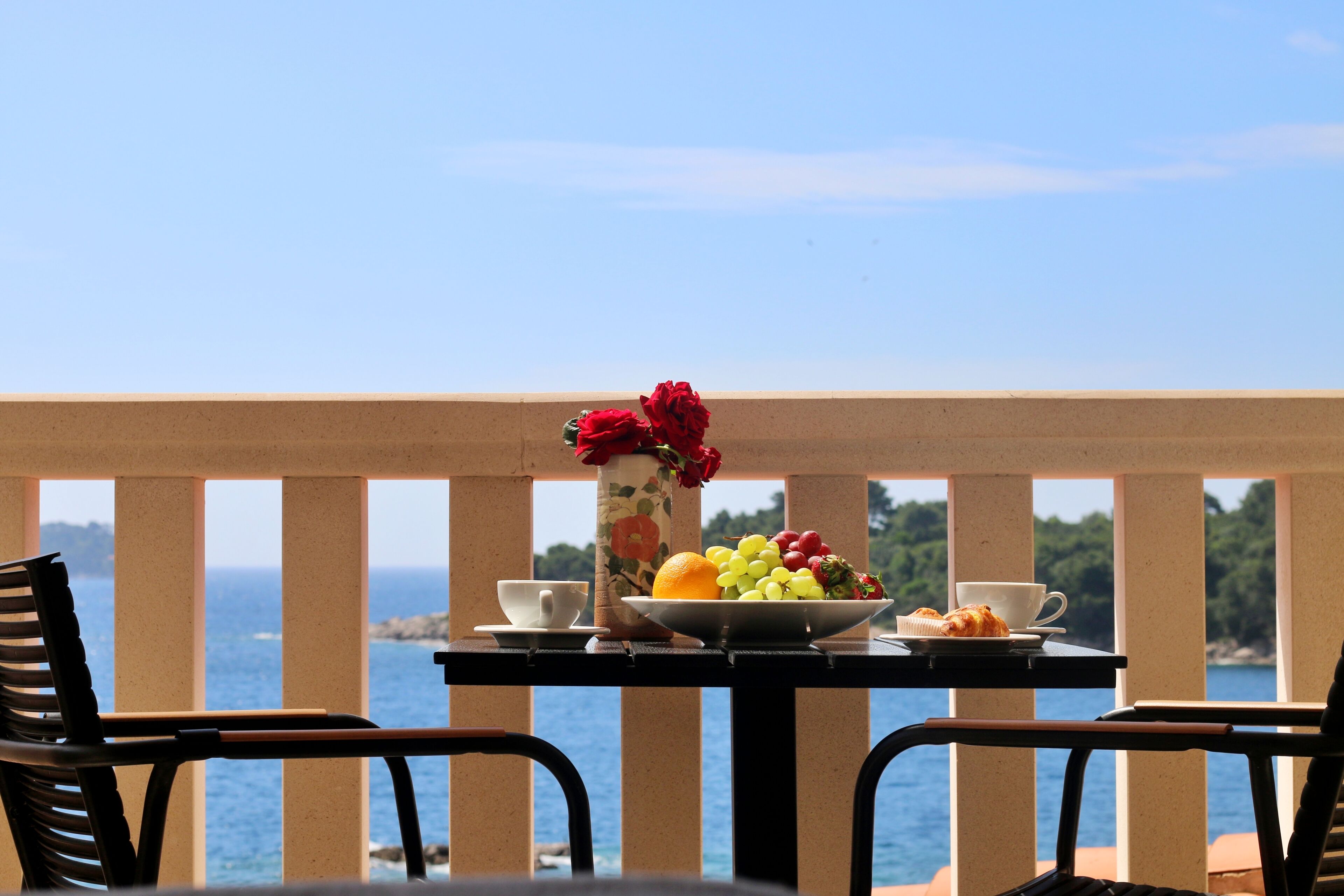 A table with a fruit bowl, a vase with flowers, and a croissant on a balcony overlooking the sea.