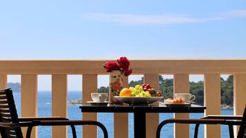 A table with a fruit bowl, a vase with flowers, and a croissant on a balcony overlooking the sea.