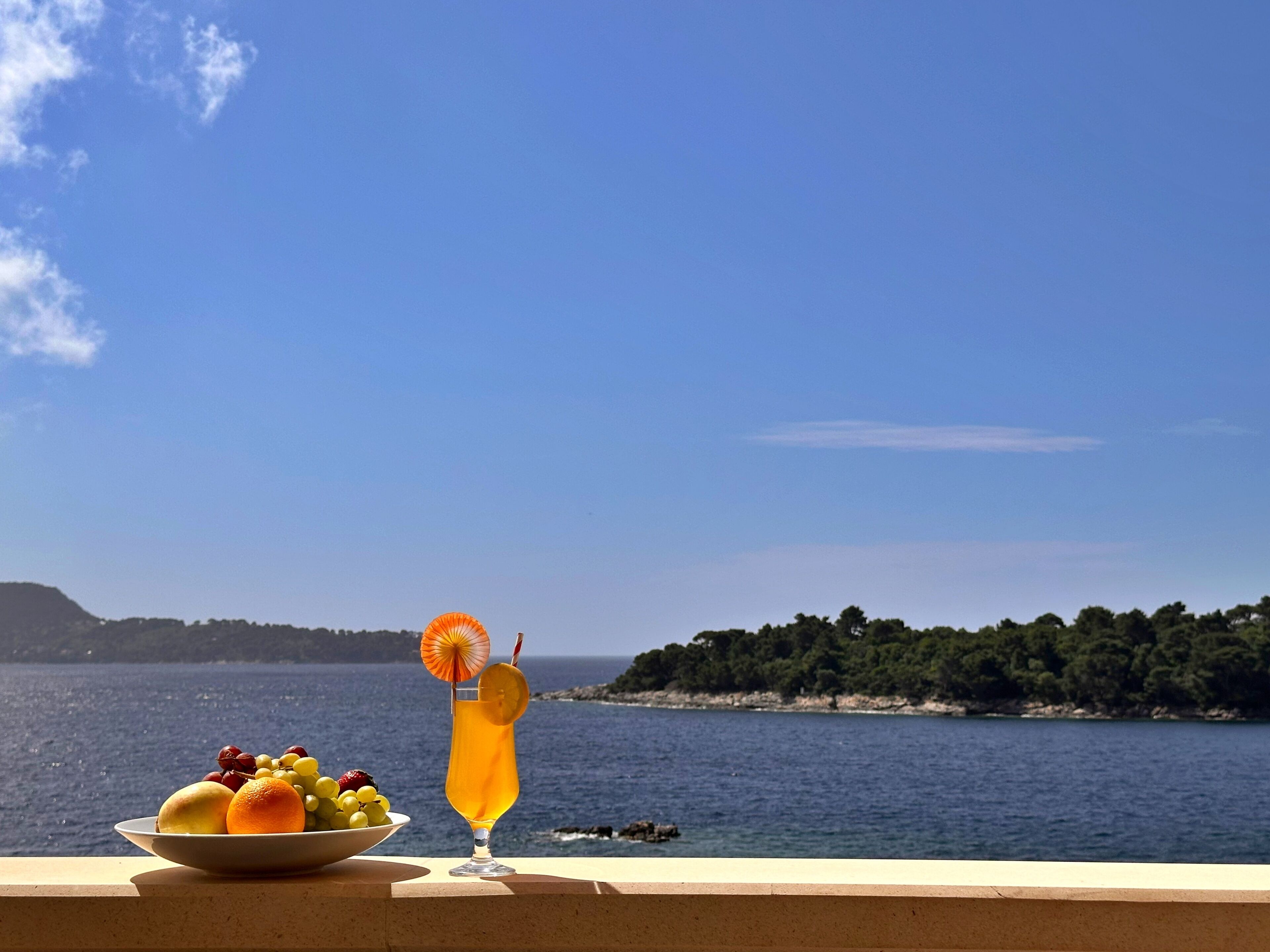 A balcony with a fruit bowl and a drink, overlooking a body of water and a distant shoreline.