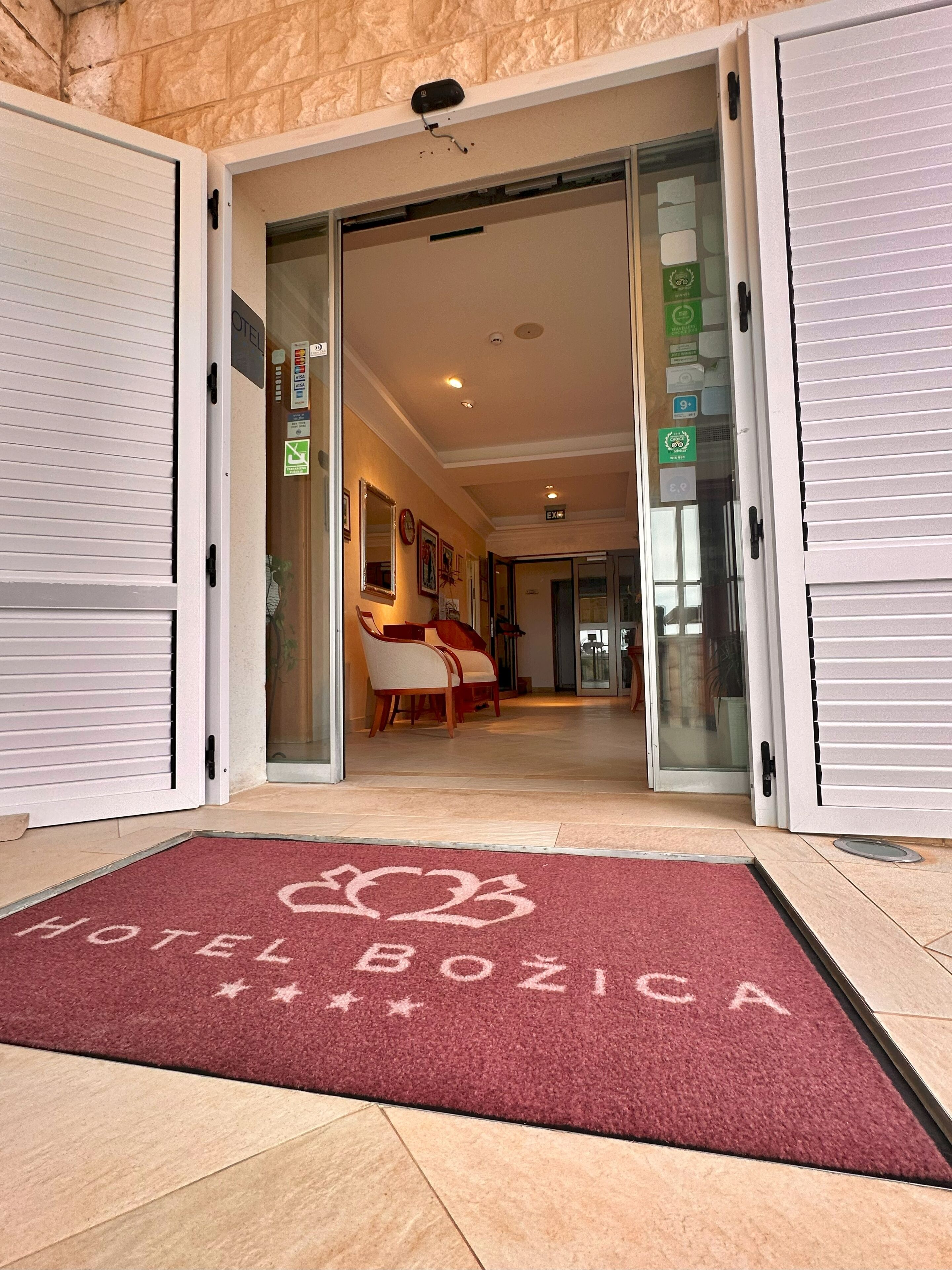 L’entrée d’un hôtel, avec un paillasson rouge sur lequel on peut lire « HÔTEL BOZICA », un espace de réception avec des chaises et un panneau indiquant clairement la sortie.