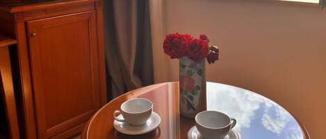 A room with a wooden cabinet, a TV, a glass table with two cups and a vase of red roses, and a window with a view of a body of water and mountains.