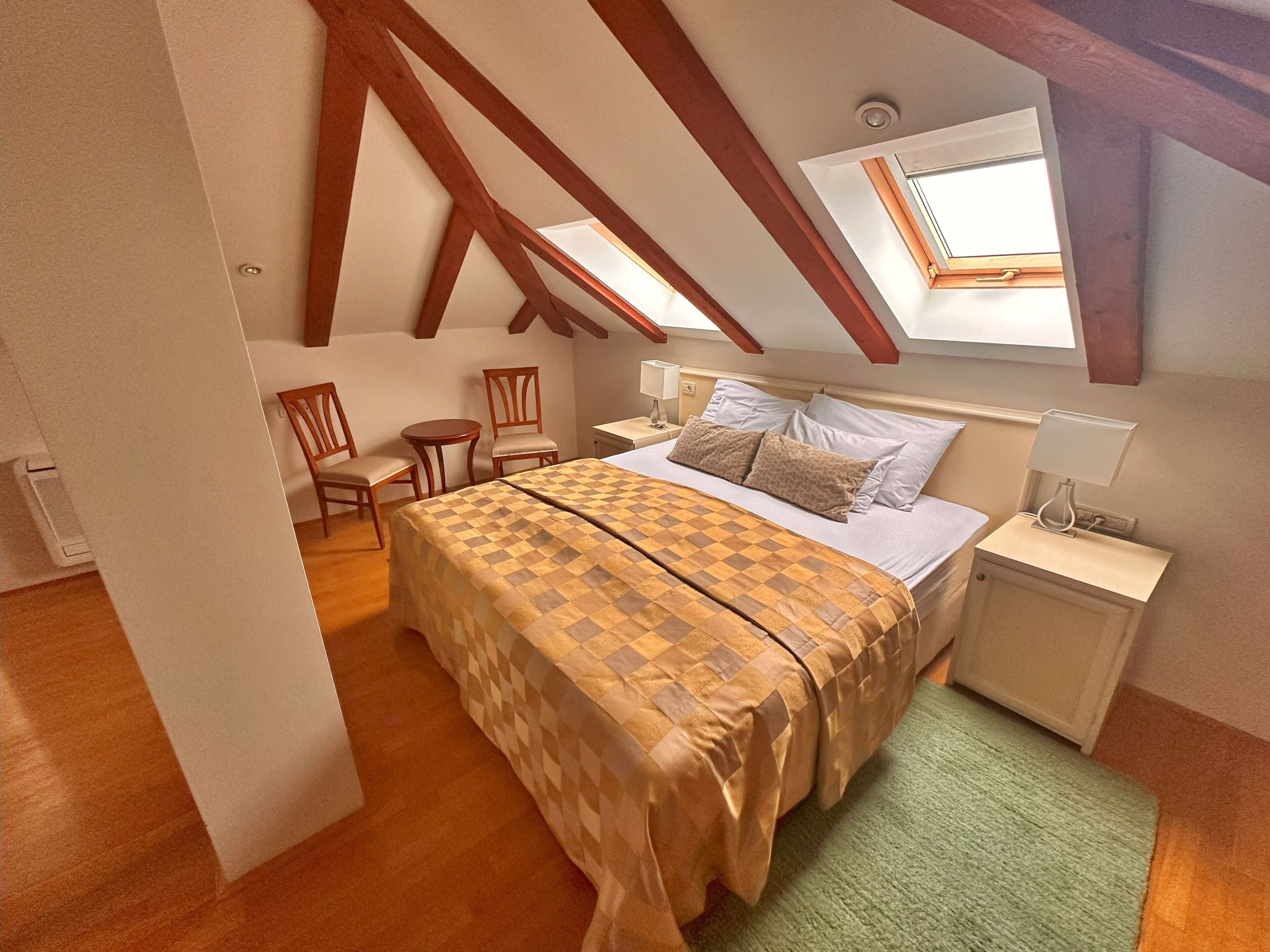 A bedroom with a high ceiling, wooden beams, a bed with a patterned bedspread, a bedside table with a lamp, and a window with a view.
