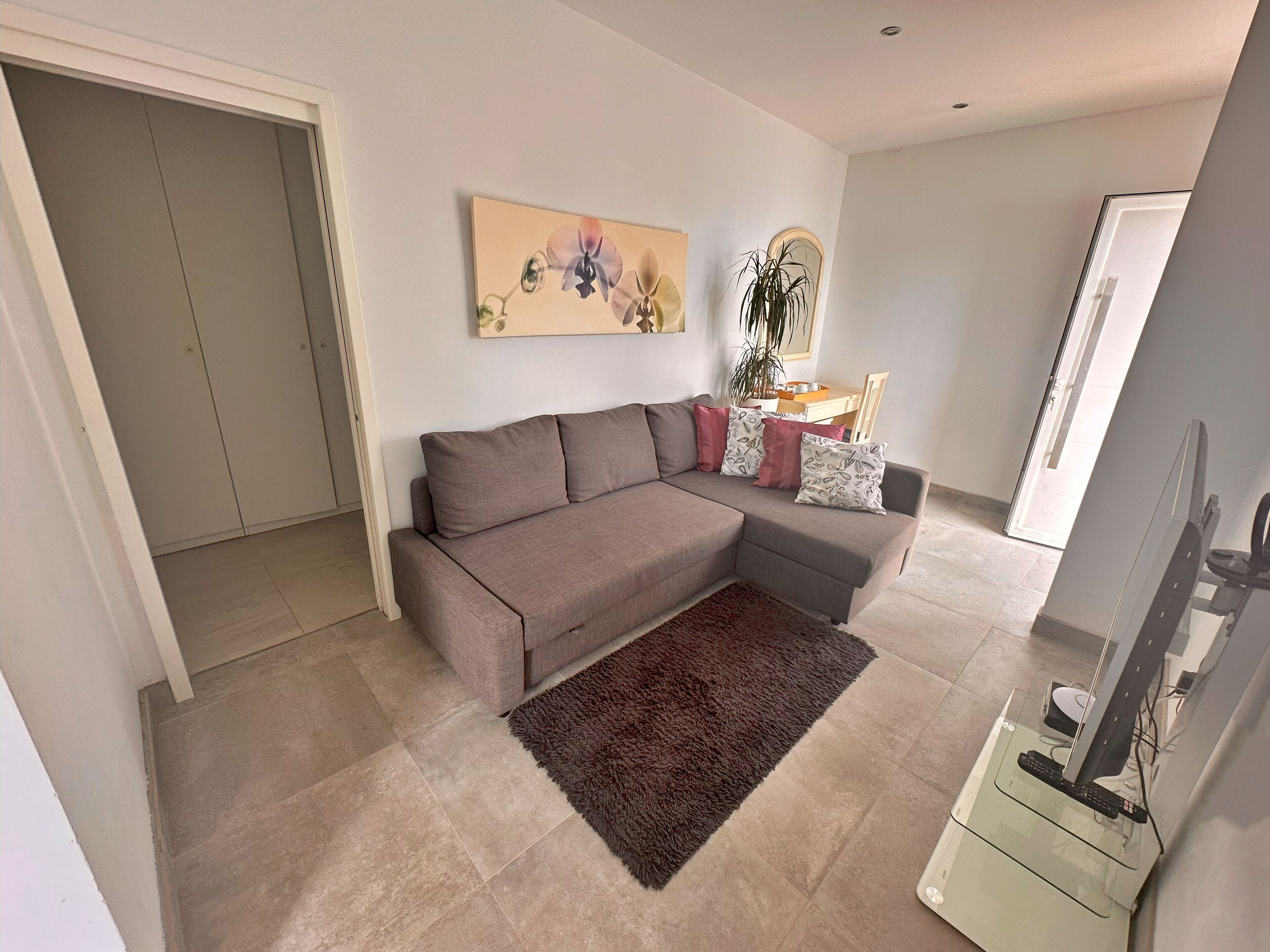 Una sala moderna con un sofá seccional gris, un tapete marrón, un televisor de pantalla plana y una zona de comedor con una mesa y sillas.