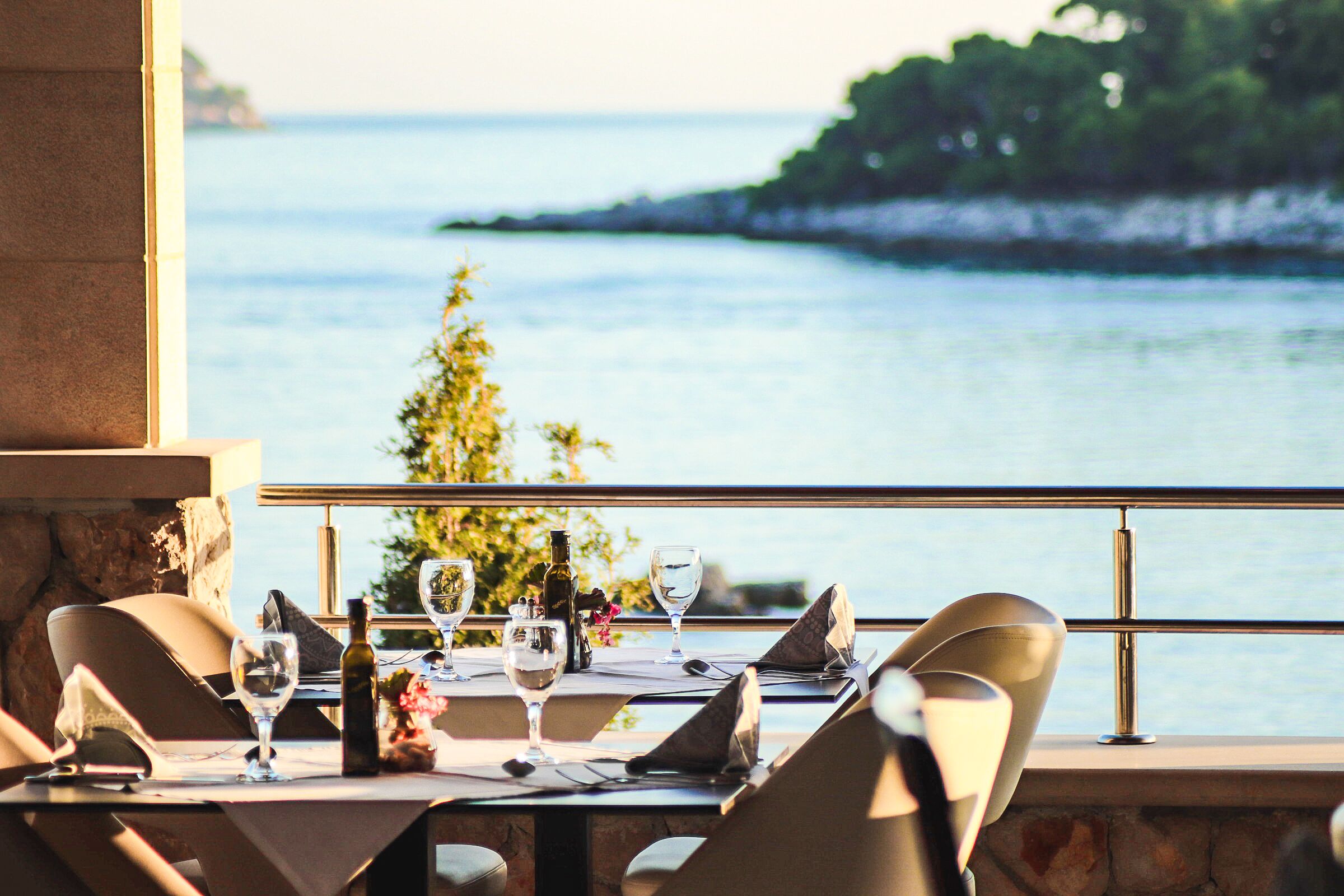 A dining table set with wine glasses, a bottle, and a flower arrangement, overlooking a body of water and greenery.
