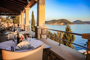 A seaside restaurant with outdoor seating overlooking a calm sea and hills. Tables are set with white tablecloths, wine glasses, and cutlery. - Hotel Bozica (Dubrovnik)