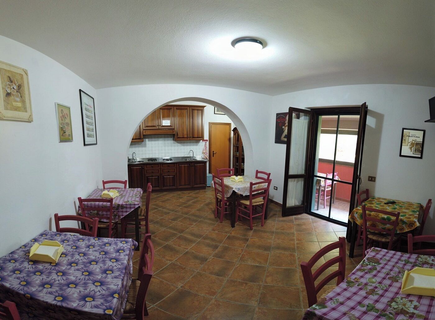 A dining area with four tables, each with a floral tablecloth. There are two wooden chairs around each table. The room has a tiled floor and white walls. An archway leads to a kitchen with wooden cabinets and a tiled backsplash.