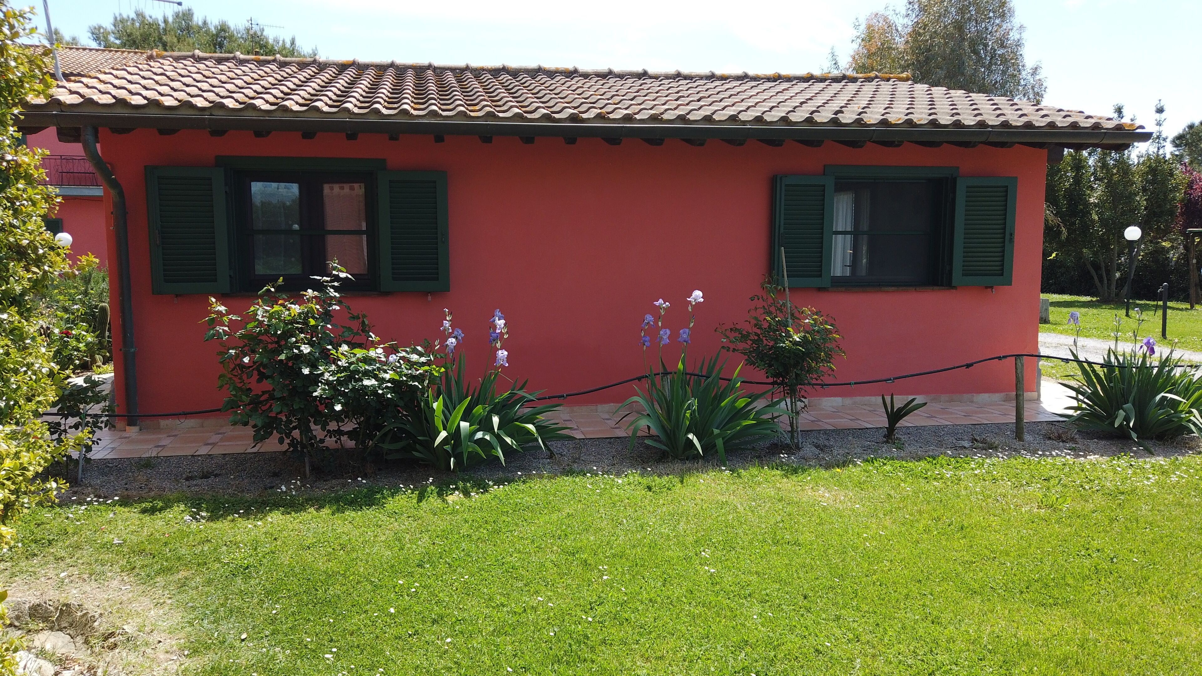 Un bâtiment rouge aux volets verts, entouré d’un jardin où poussent différentes plantes et fleurs.