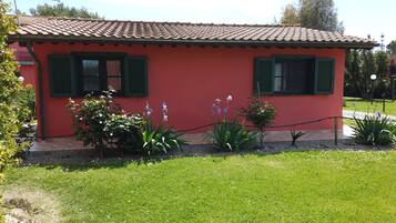 Un bâtiment rouge aux volets verts, entouré d’un jardin où poussent différentes plantes et fleurs.