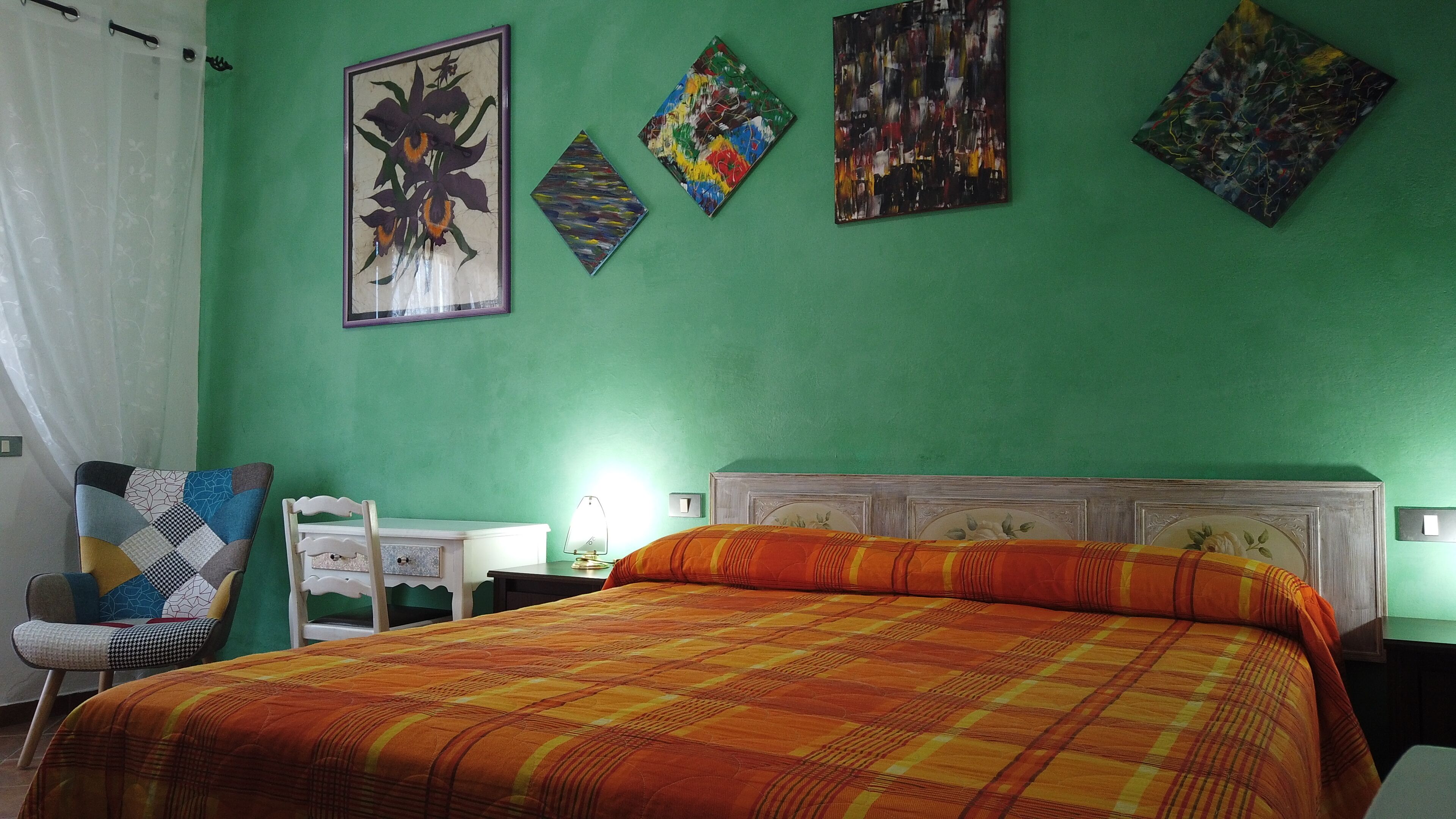 Una habitación con pared verde, una cama con colcha de cuadros naranja y amarillo, cabecera blanca y una silla con cojín de diseño geométrico. Hay cuatro cuadros en la pared y una mesita de noche con lámpara.