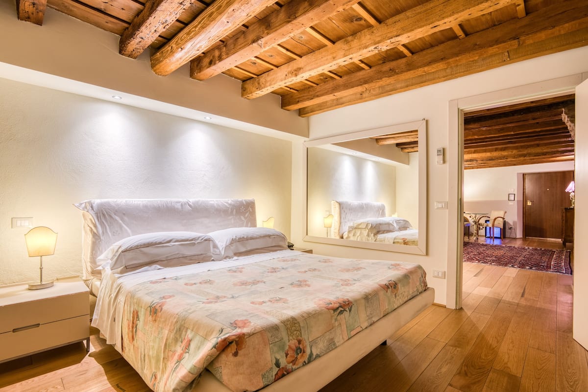 A bedroom with a wooden ceiling, a large bed with floral bedding, a nightstand with a lamp, and a mirror reflecting the room.