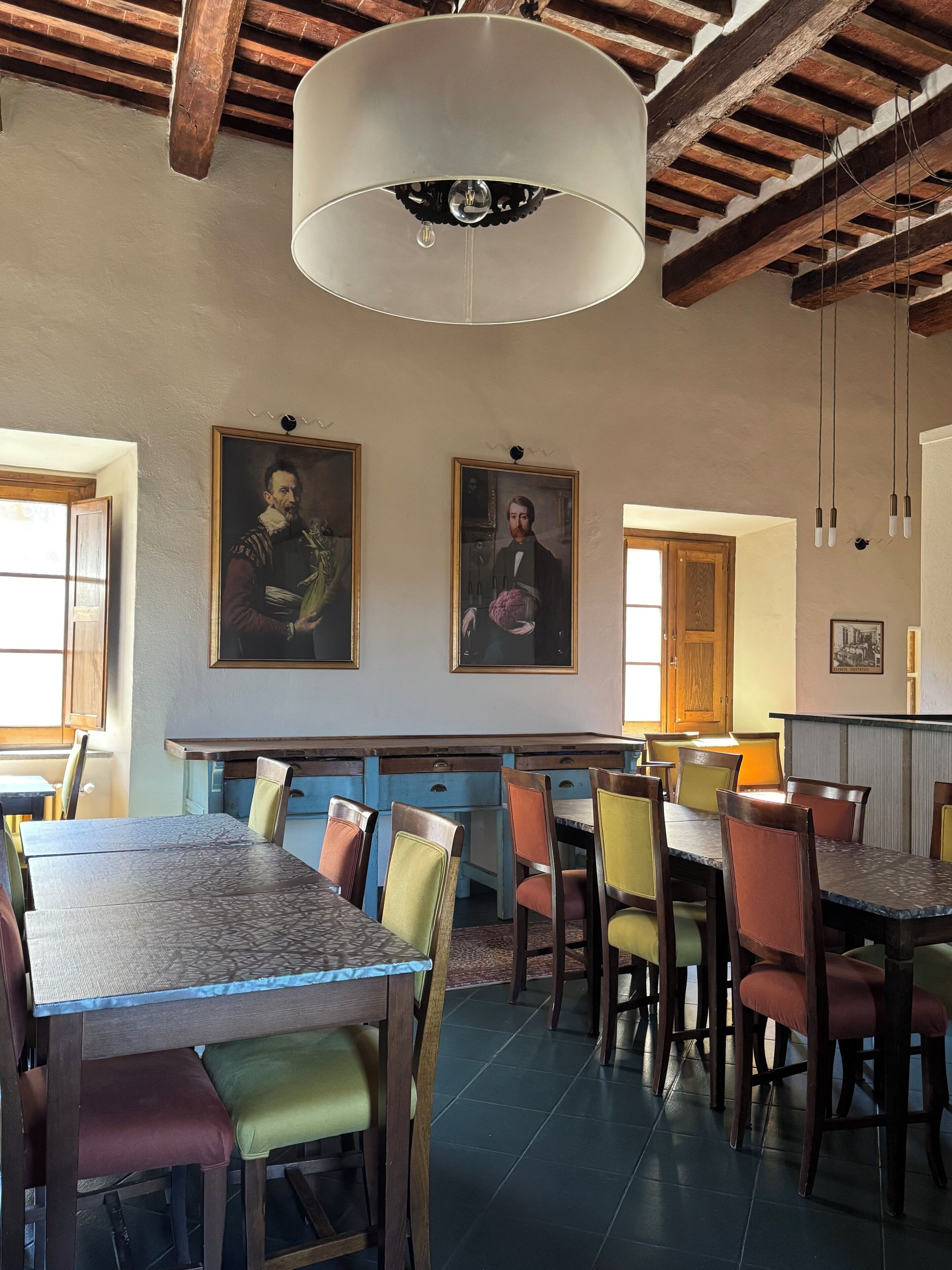 Un comedor con mesas y sillas de madera, una lámpara colgante y dos retratos enmarcados en la pared.