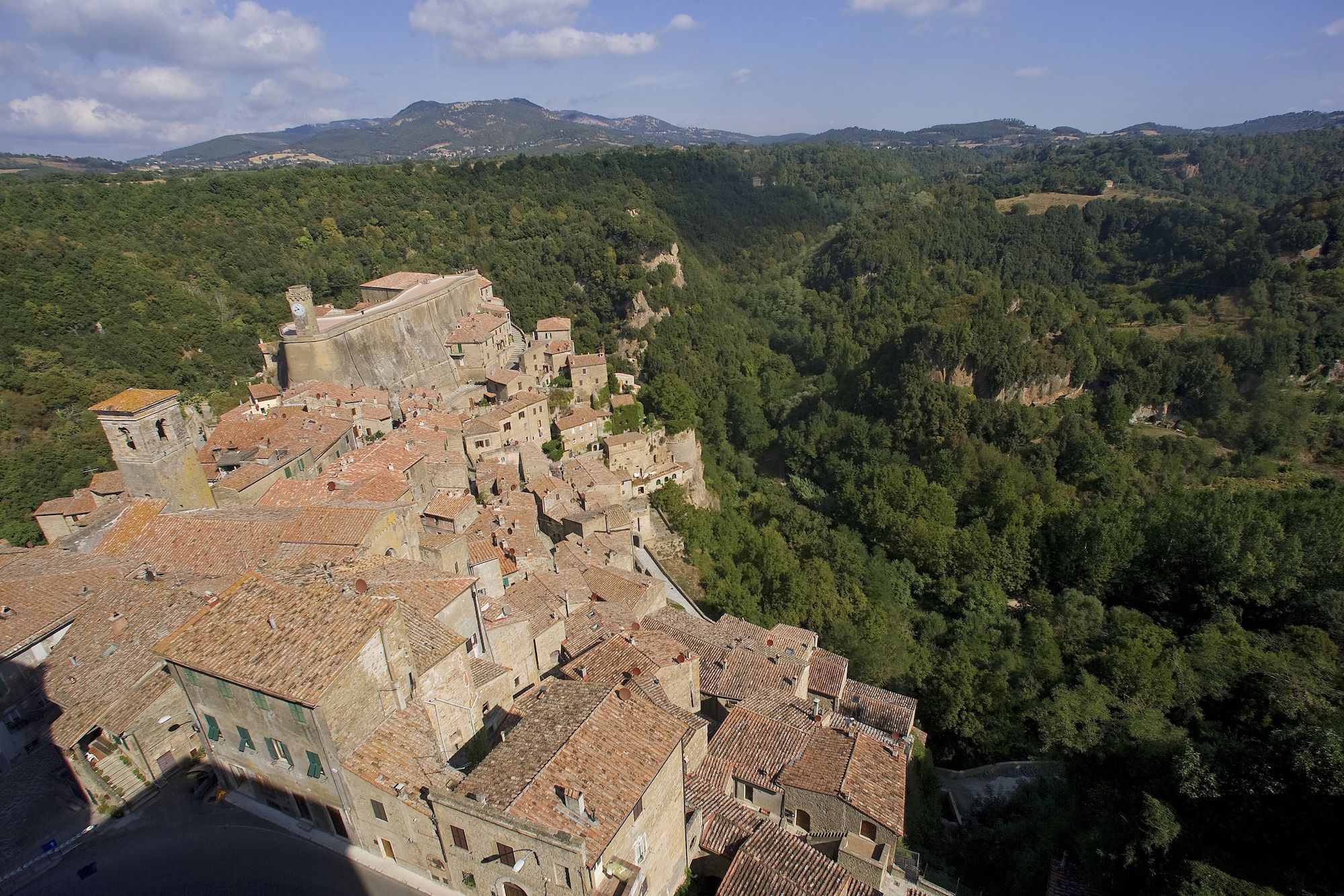 A historic town with terracotta-roofed buildings nestled in a valley, surrounded by dense forests and hills.