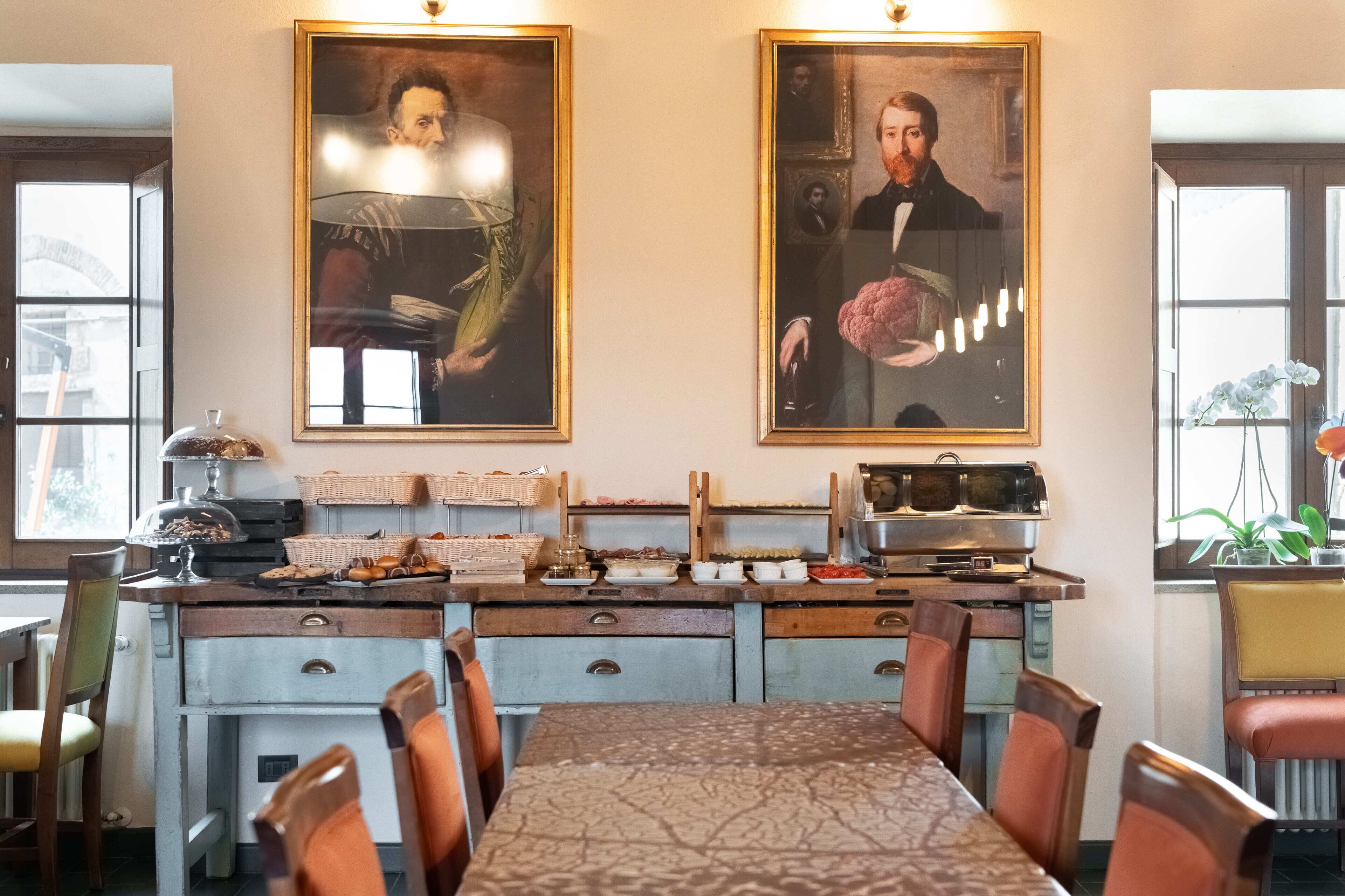 Un comedor con una mesa larga, sillas y un sistema de buffet con diversos alimentos. Hay dos retratos grandes enmarcados en la pared.