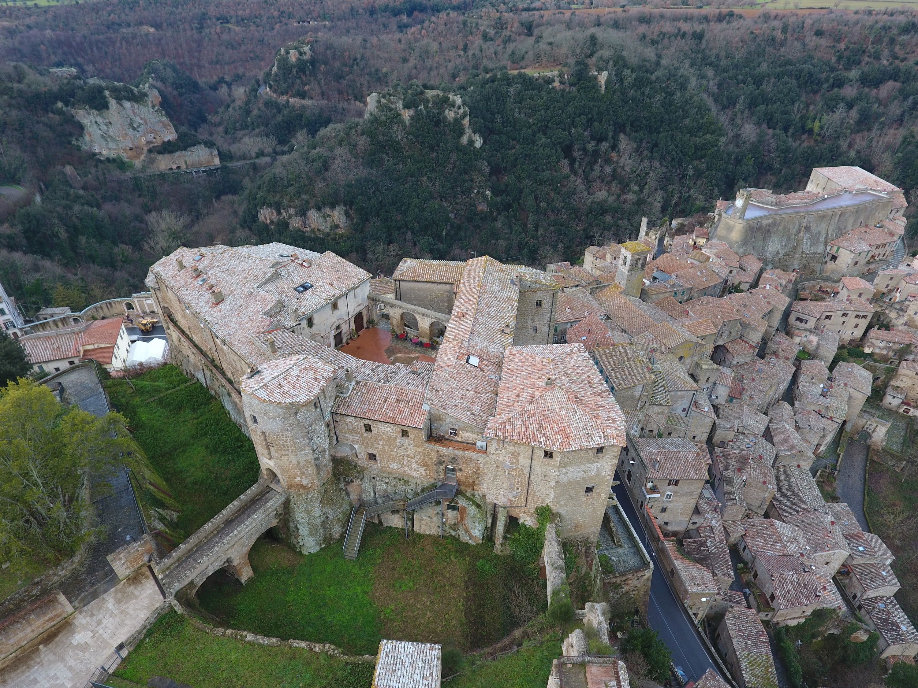An aerial view of a historic town with medieval architecture, including a prominent castle-like structure with a courtyard and a bridge.