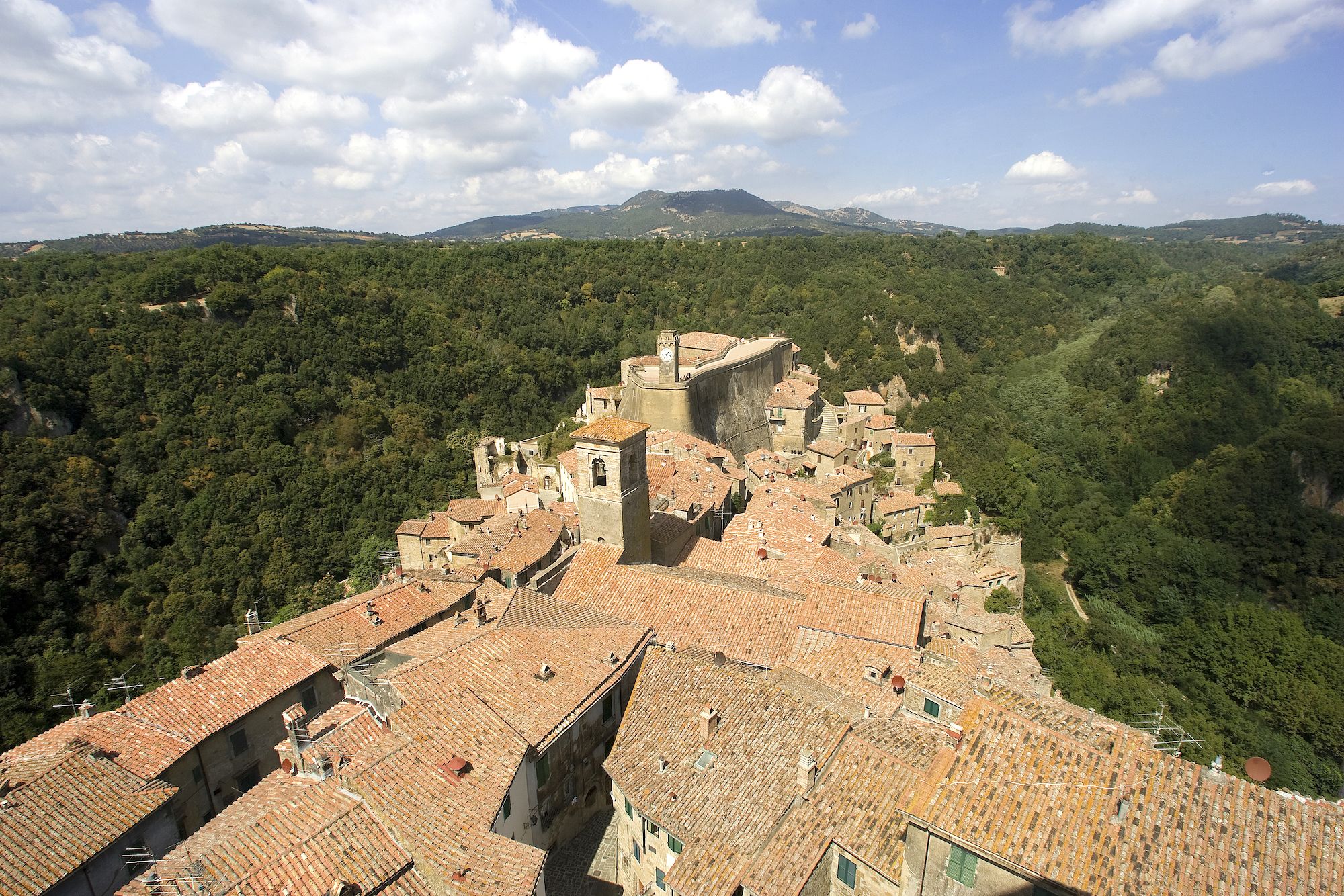 A historic town with terracotta-roofed buildings, a prominent tower, and a lush green valley in the background.