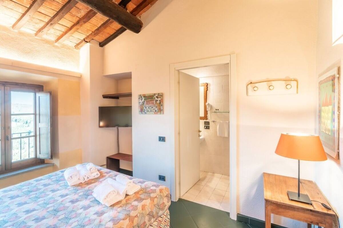 A bedroom with a bed, a wooden table with a lamp, a bathroom door, and a wall-mounted light fixture.