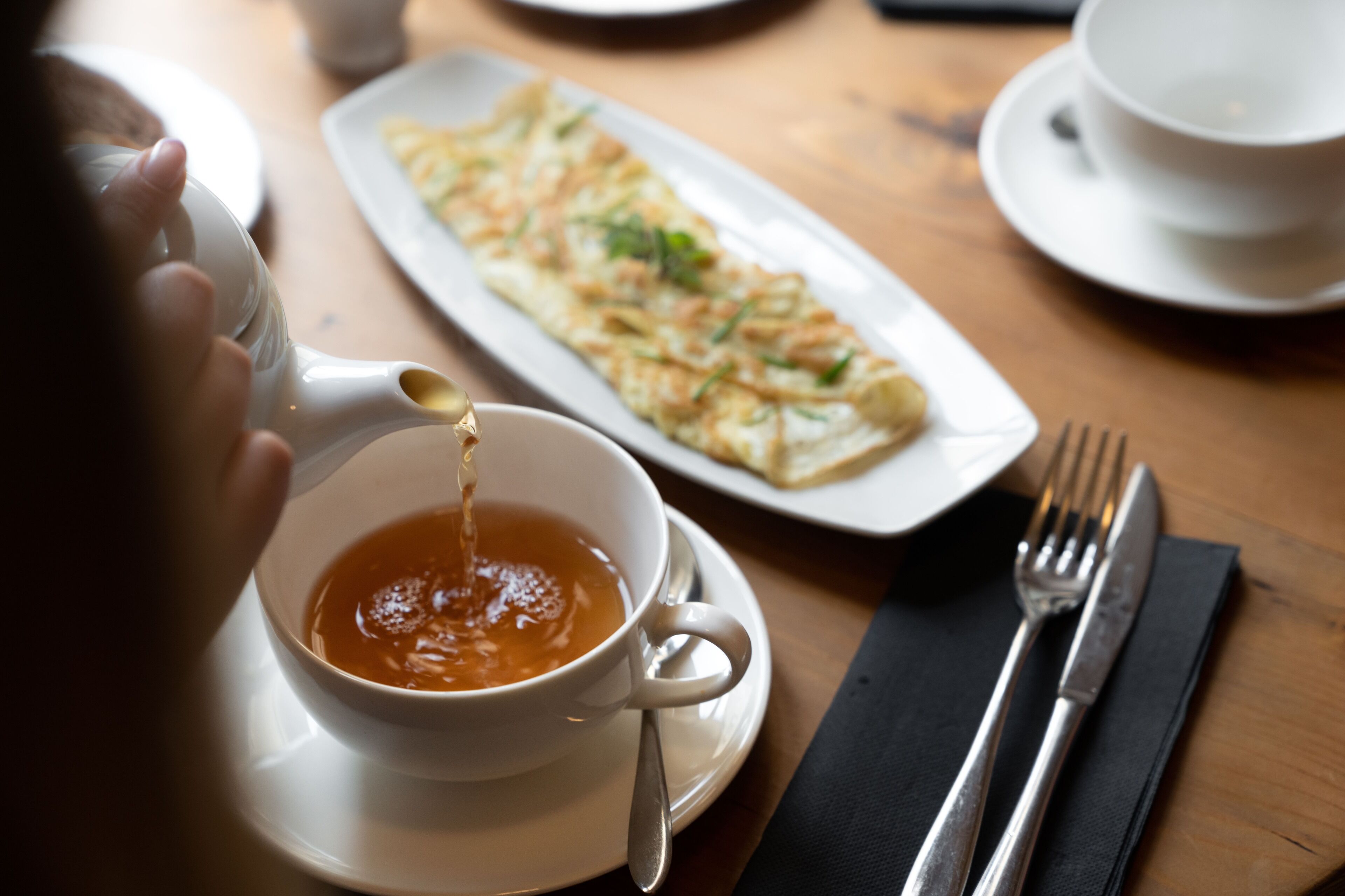 A dining table with a white teapot pouring tea into a cup, a plate of food, and cutlery.
