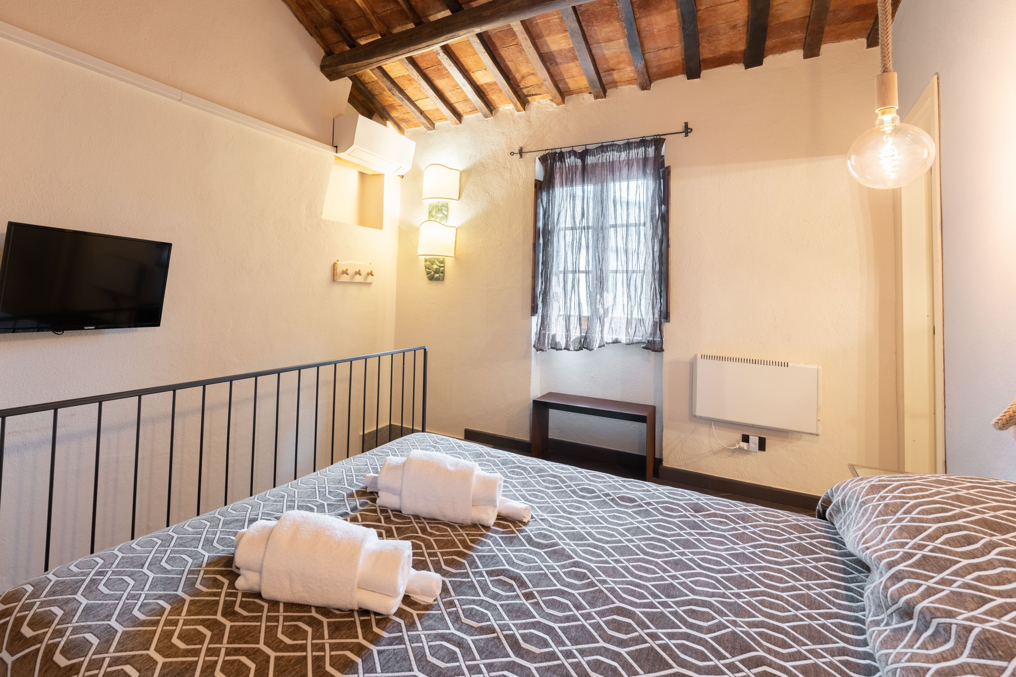 Una habitación de hotel con cama, televisor, ventana con cortinas y techo con vigas a la vista.