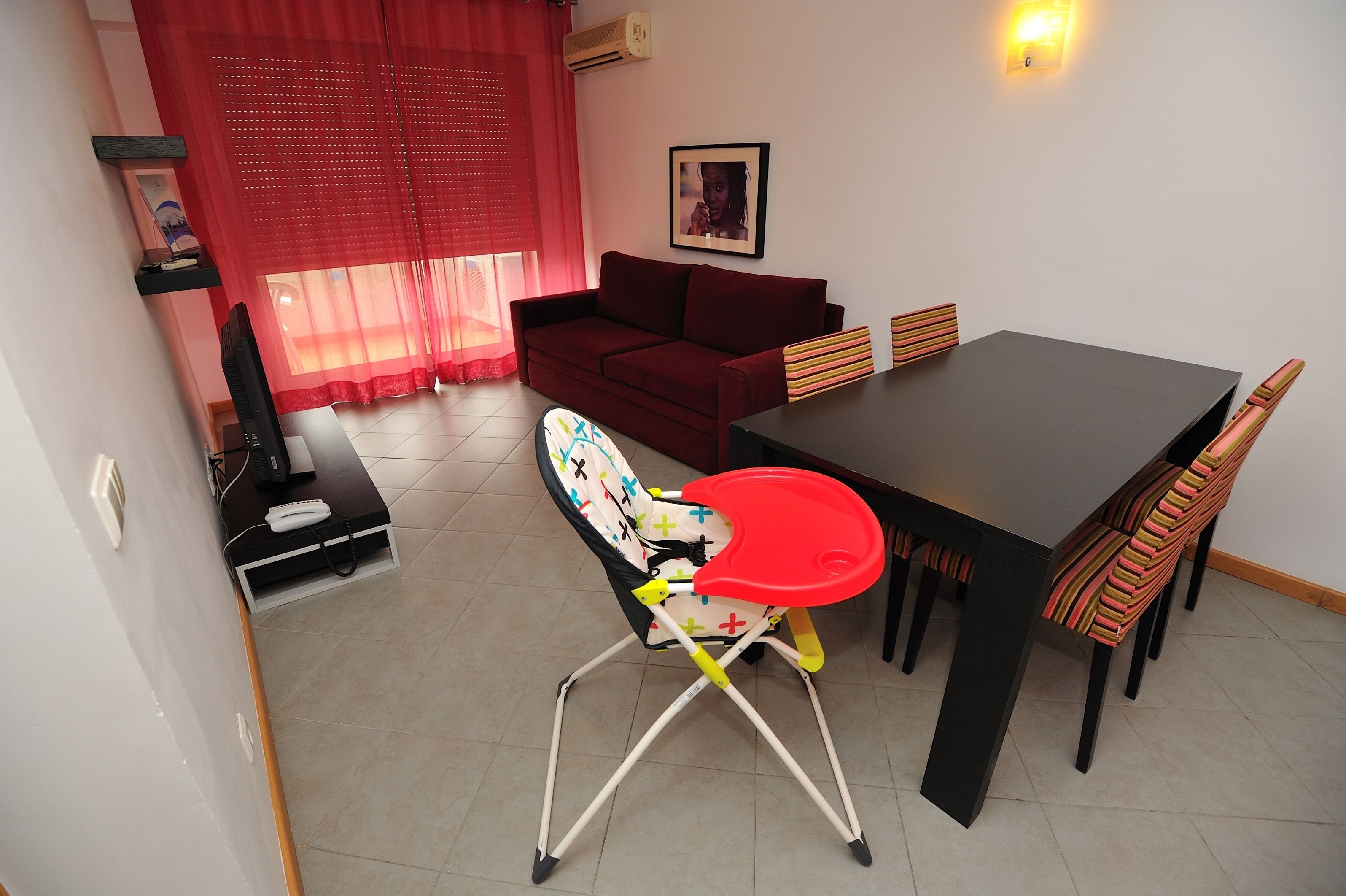 A dining area with a table, chairs, a red chair with a tray, a sofa, and a red curtain.