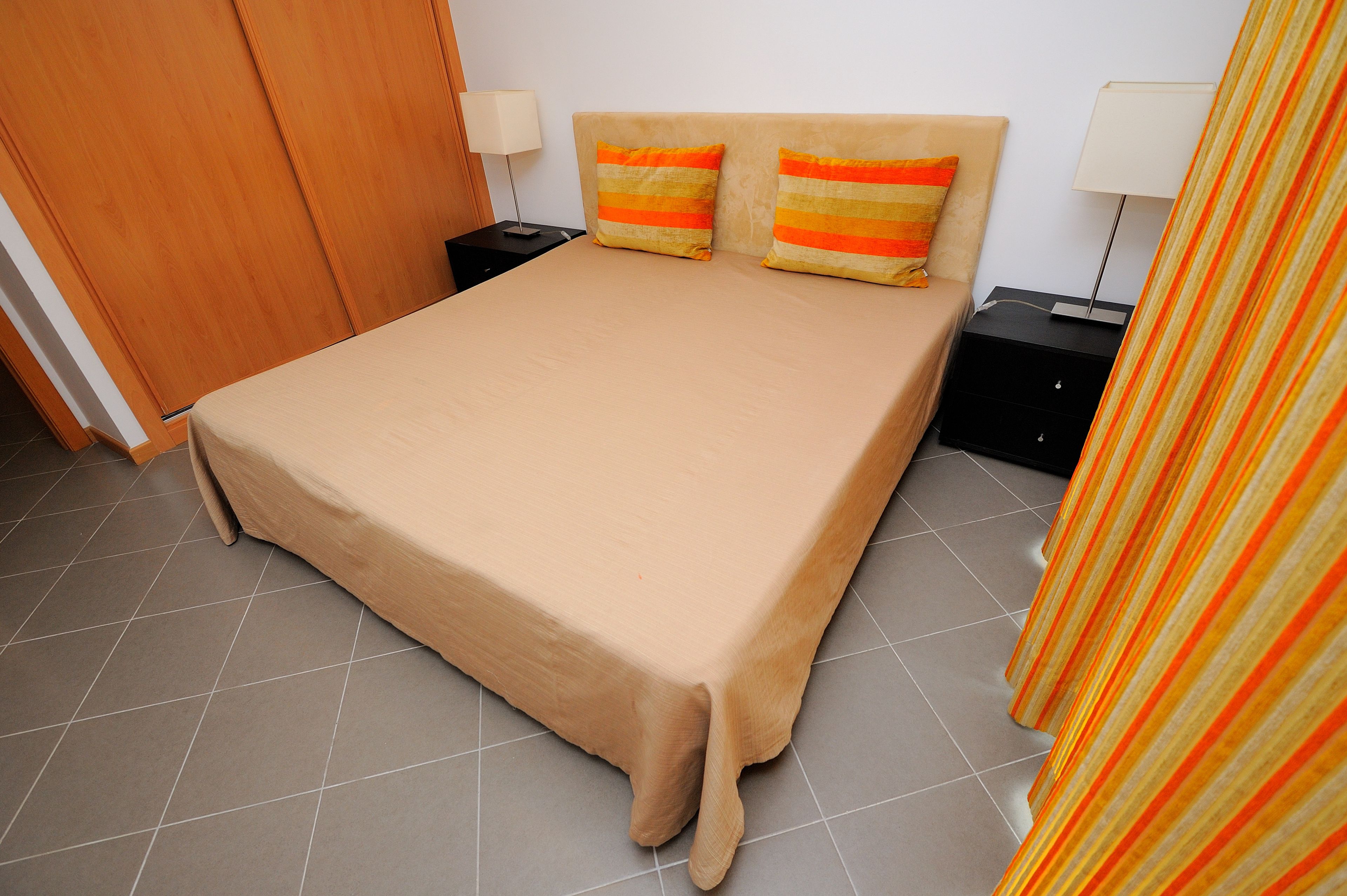 Un lit bien fait, recouvert d’une couverture beige, avec deux oreillers à rayures orange et jaunes, deux tables de chevet avec des lampes et un rideau à rayures.