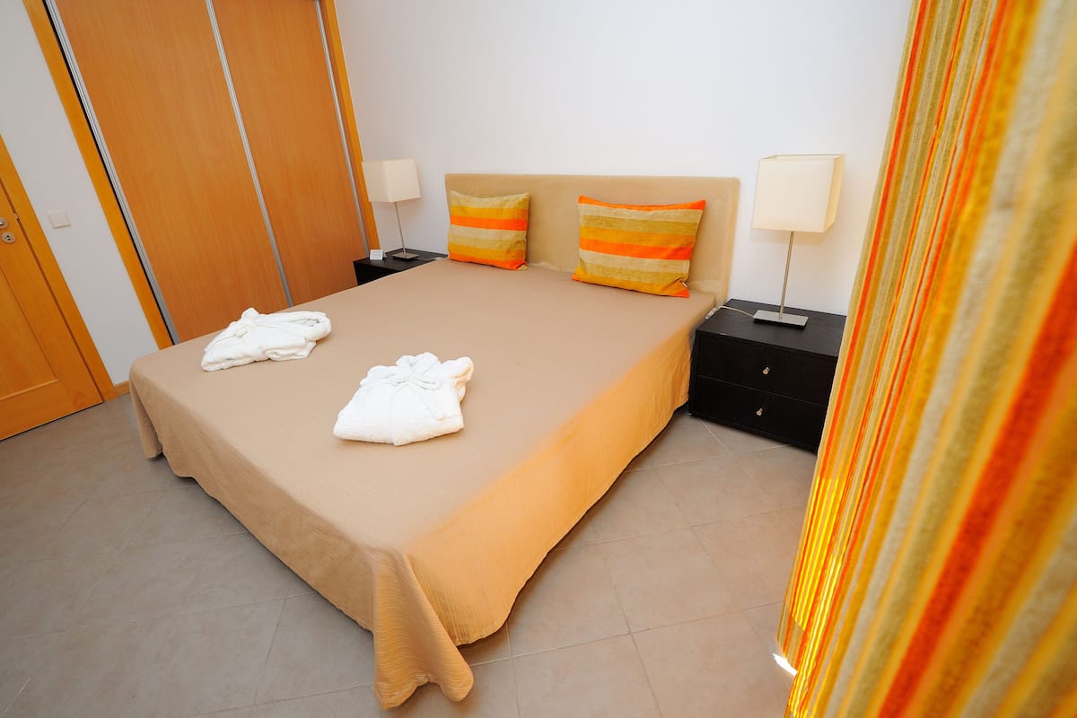 Un letto rifatto con due asciugamani bianchi, una coperta beige e due cuscini a righe. Sono presenti due comodini con lampade e un armadio in legno sullo sfondo.