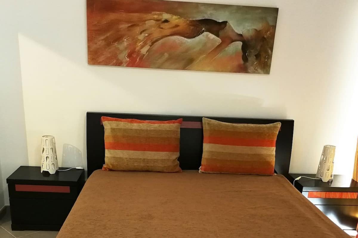 Un letto con una coperta marrone, due cuscini a righe, un quadro sopra il letto e un comodino con una lampada e un caricabatterie per telefono.