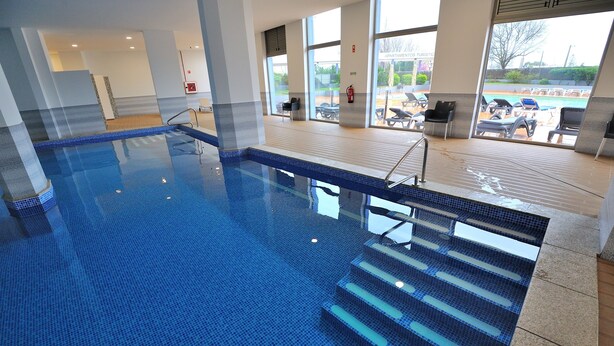 An indoor swimming pool with clear blue water, steps leading into the pool, and a view of the outside through large windows.