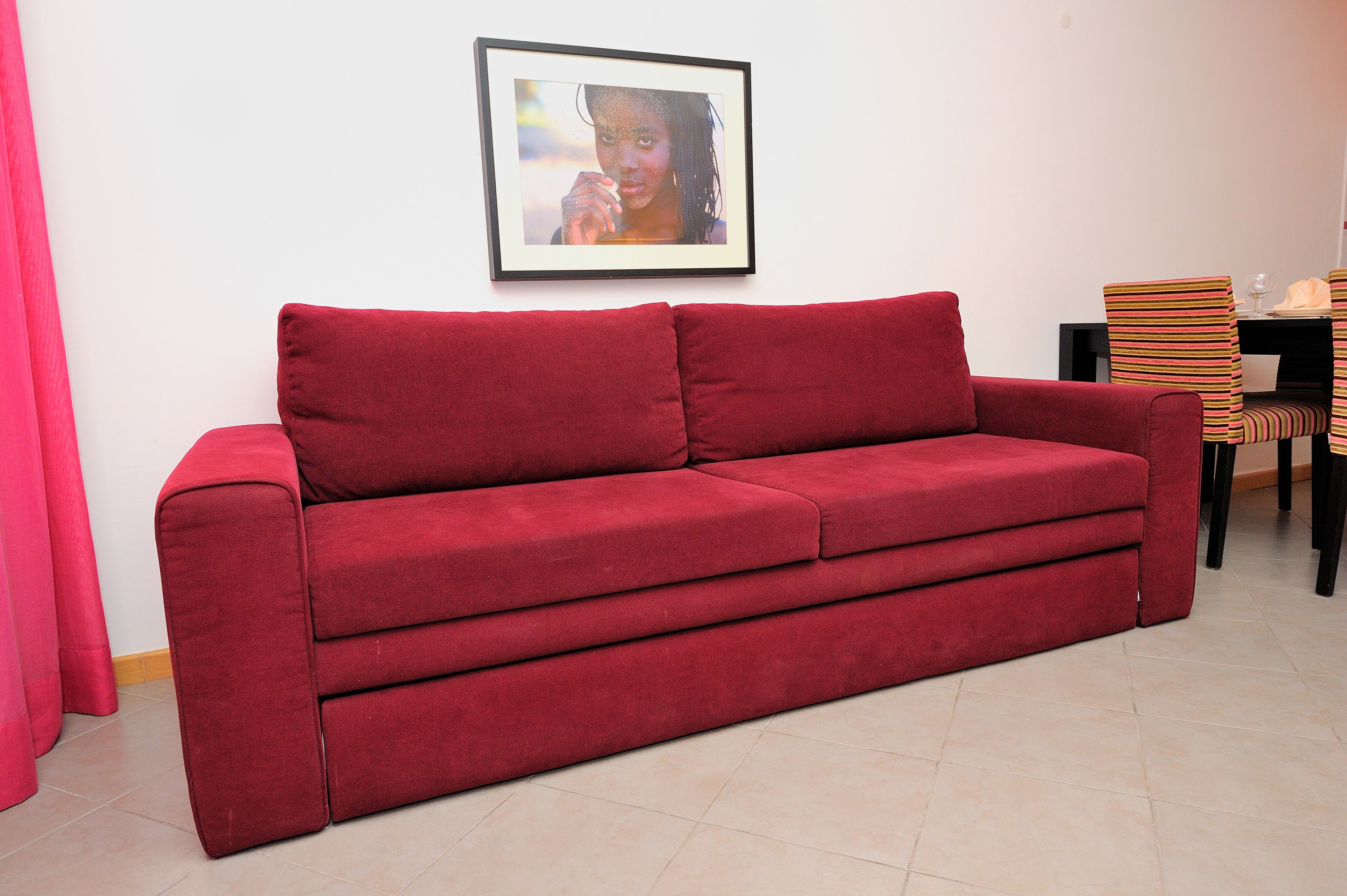 A red sofa in a room with a framed picture on the wall, a dining table with chairs, and a pink curtain.