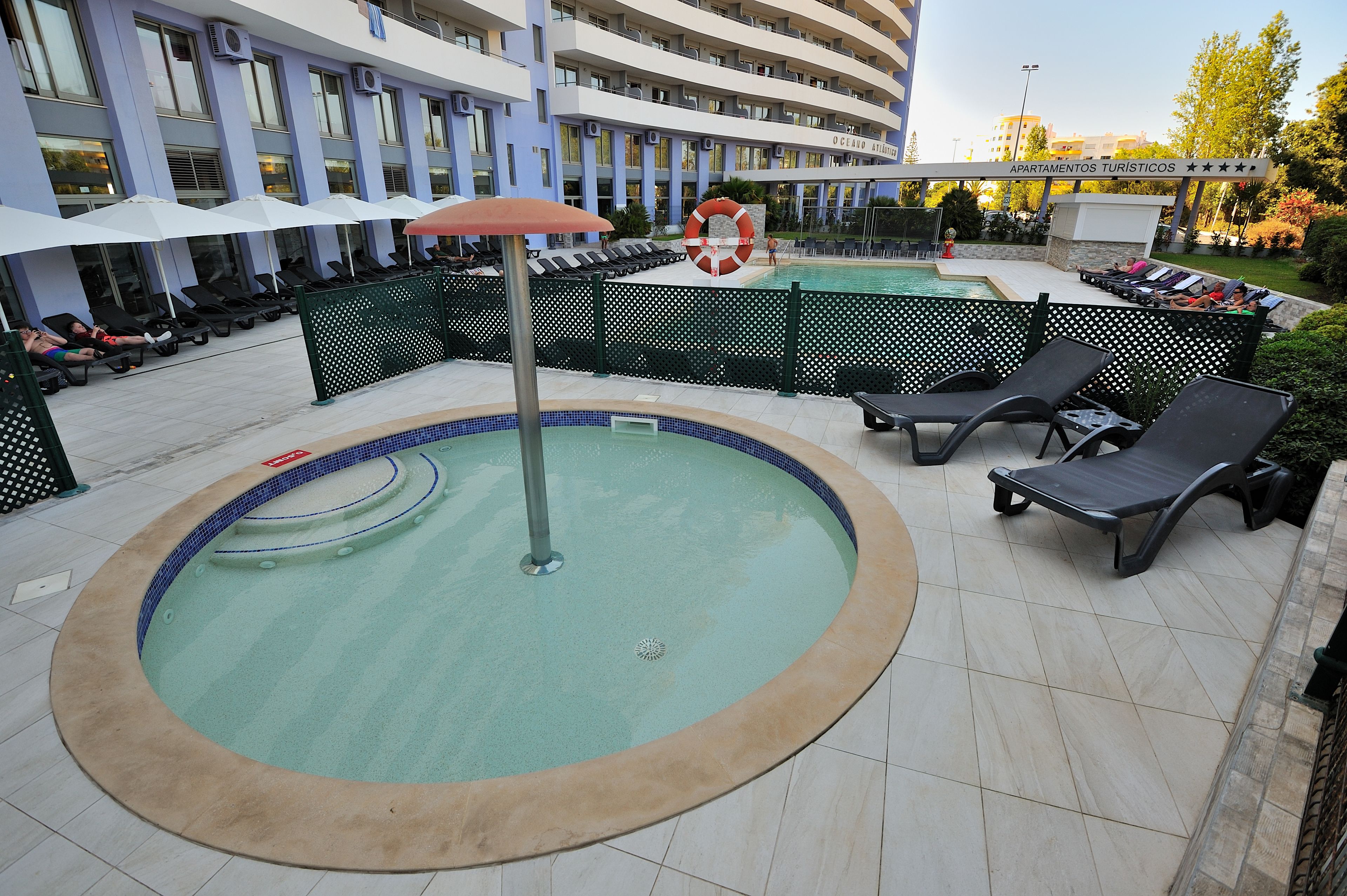 Un espace piscine avec des chaises longues et des parasols, entouré d’une clôture, et un bâtiment portant l’inscription « Apartamentos Turísticos » en arrière-plan.