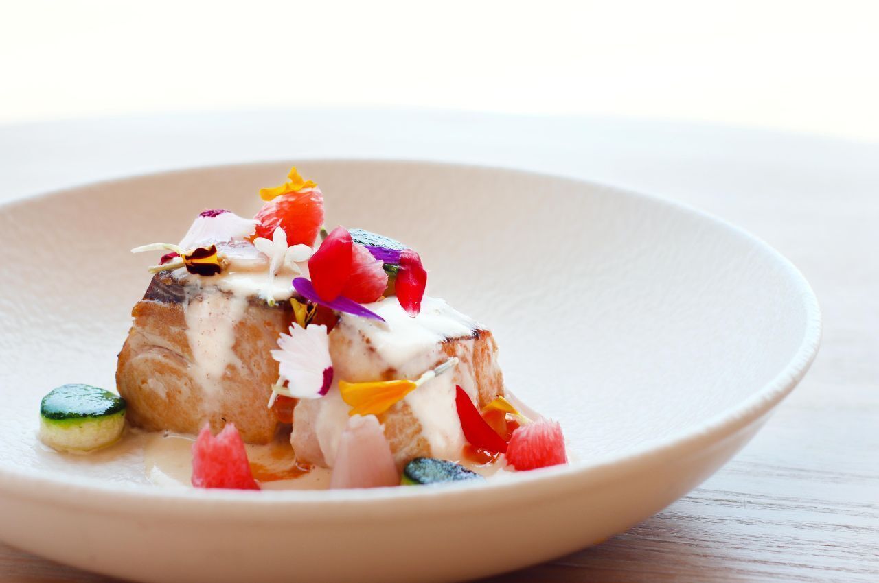 Un plato con un trozo de pescado cubierto de flores comestibles de colores y salsa cremosa.