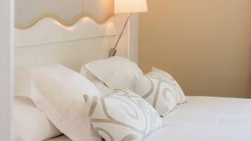 Uma cama bem arrumada com roupa de cama branca e almofadas estampadas, uma luminária fixada na parede e um desenho ondulado na cabeceira.