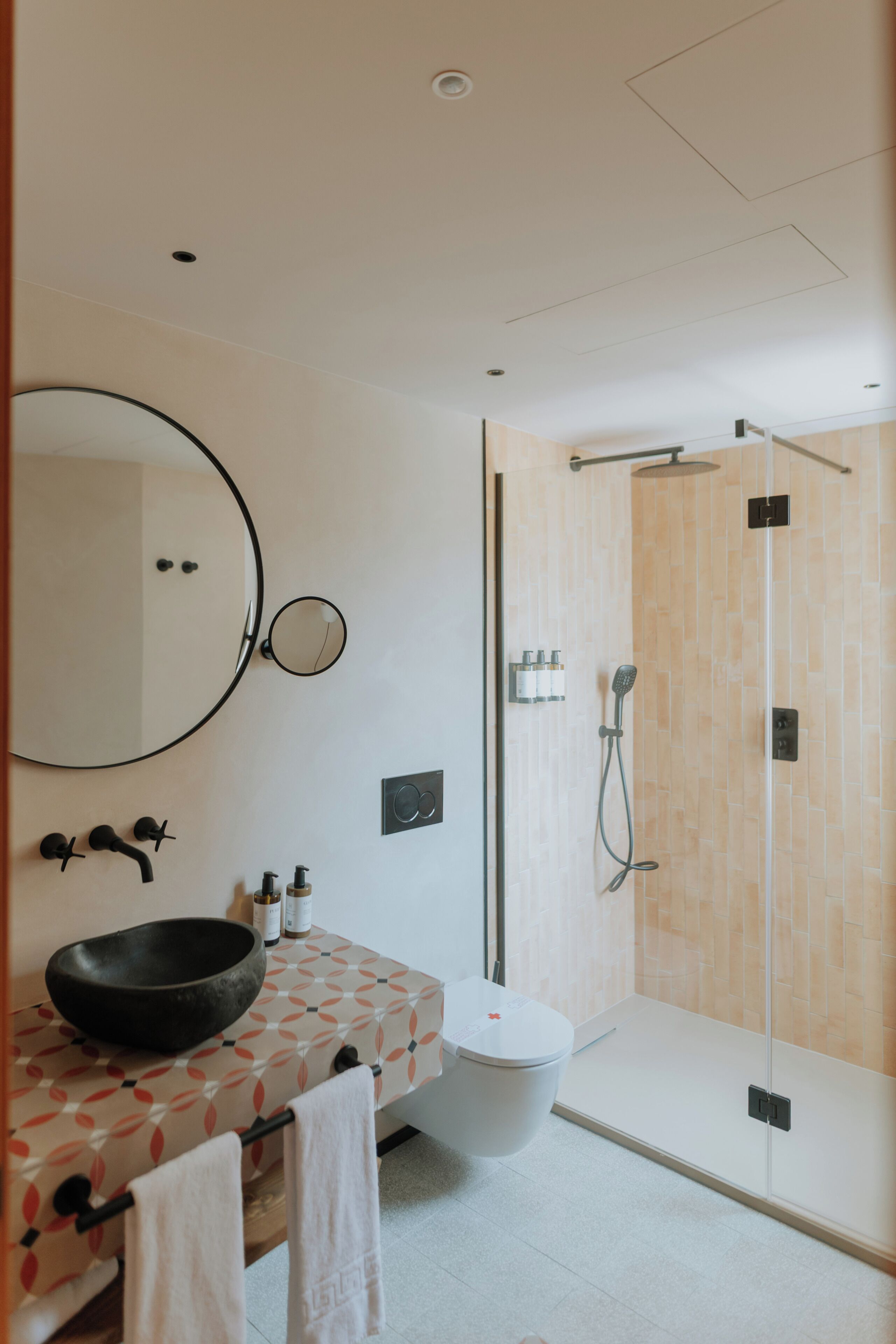 Un baño moderno con un espejo redondo, un lavabo negro, inodoro y ducha con mampara de vidrio.