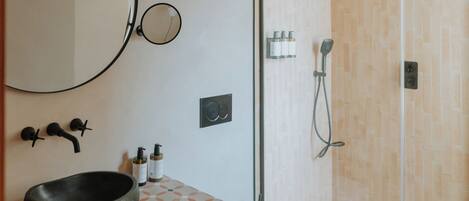 Un baño moderno con un espejo redondo, un lavabo negro, inodoro y ducha con mampara de vidrio.