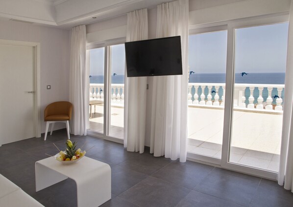 A hotel room with a large window, a flat-screen TV, a white table with a fruit bowl, and a chair.