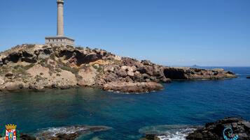 Paesaggio costiero con un faro su uno sperone roccioso, mare blu cristallino e un logo nell'angolo in basso a destra.