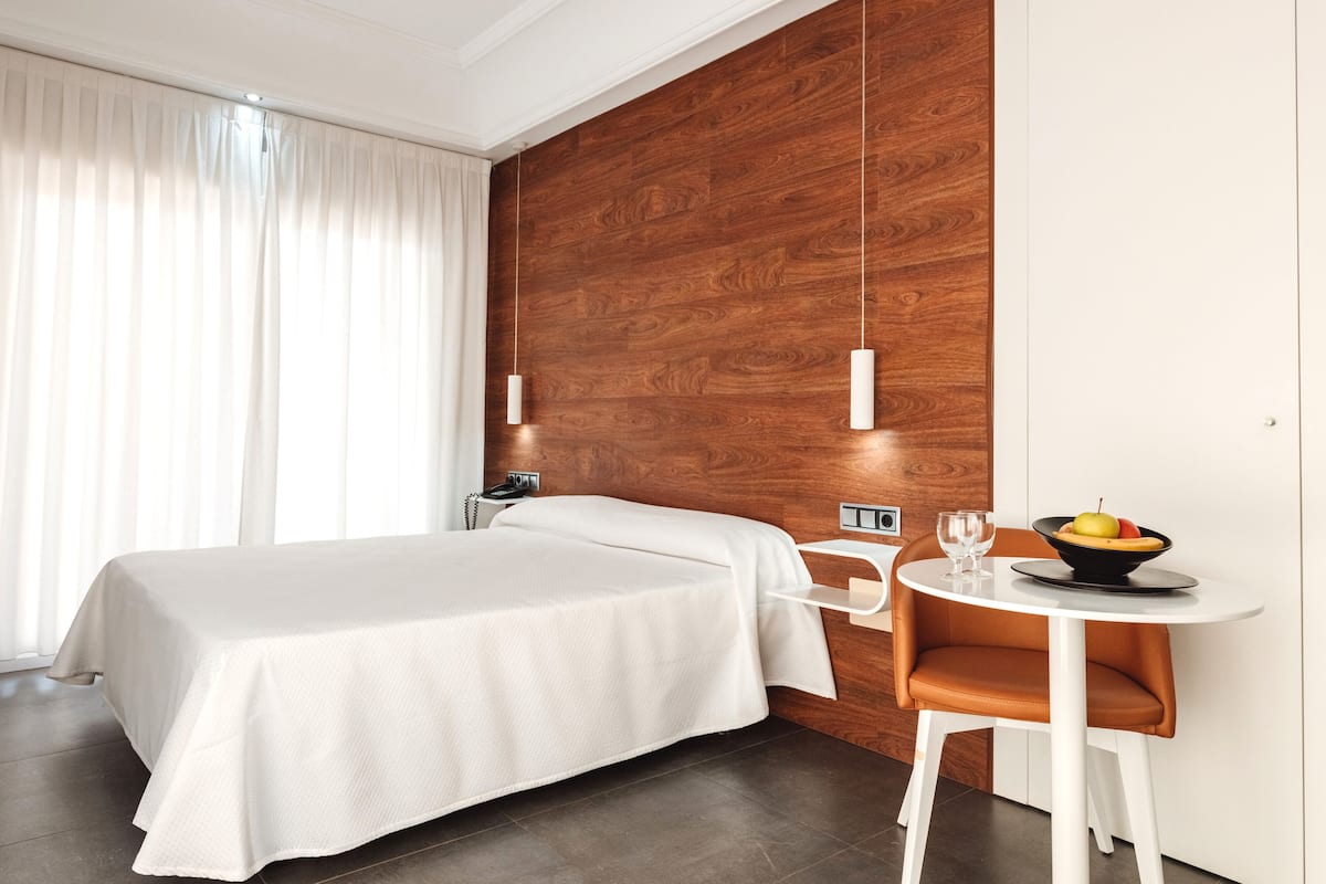 A hotel room with a bed, a small table with a bowl of fruit, a chair, and a wooden accent wall.