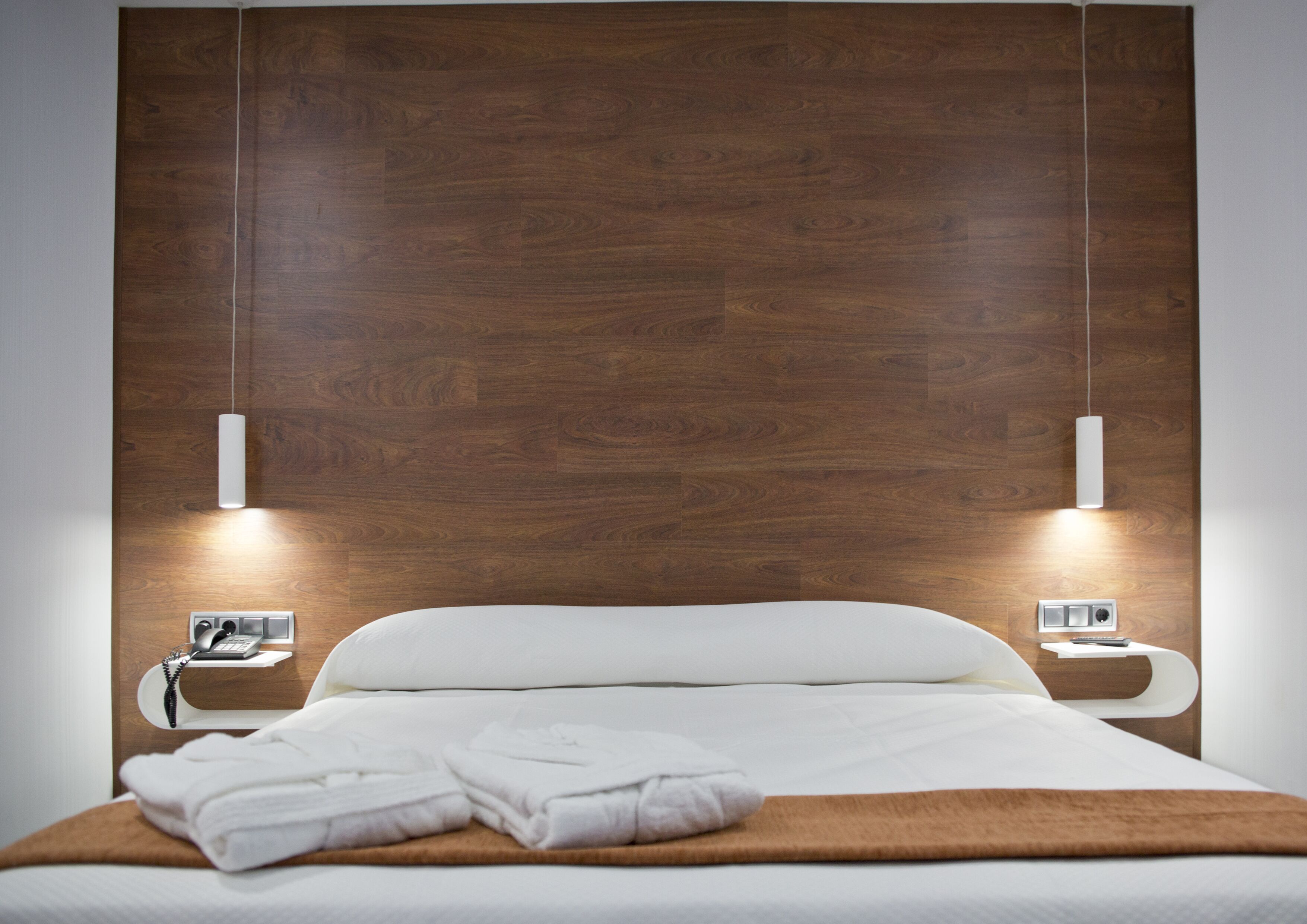 Habitación de hotel con cabecera de madera, cama con ropa de cama blanca, dos mesitas de noche con lámparas y una manta marrón sobre la cama.