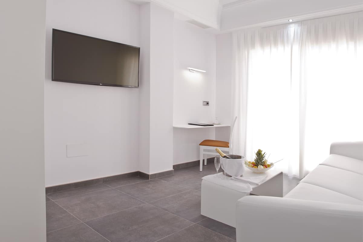 A modern living room with a flat-screen TV, a white sofa, a dining table with a chair, and a small kitchen area with a countertop and a chair.