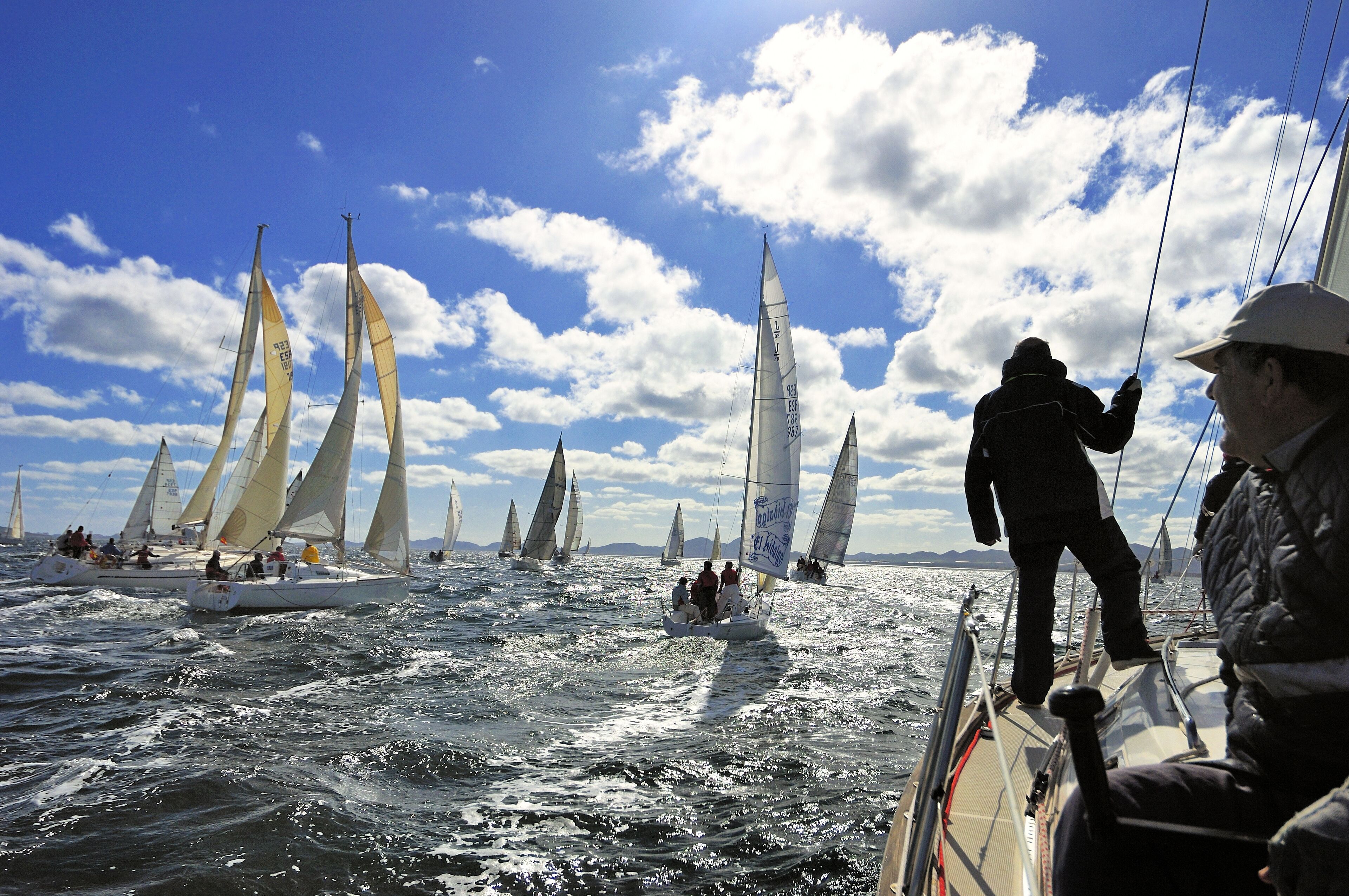 Un velero con varias velas, personas a bordo y un cielo despejado con nubes.